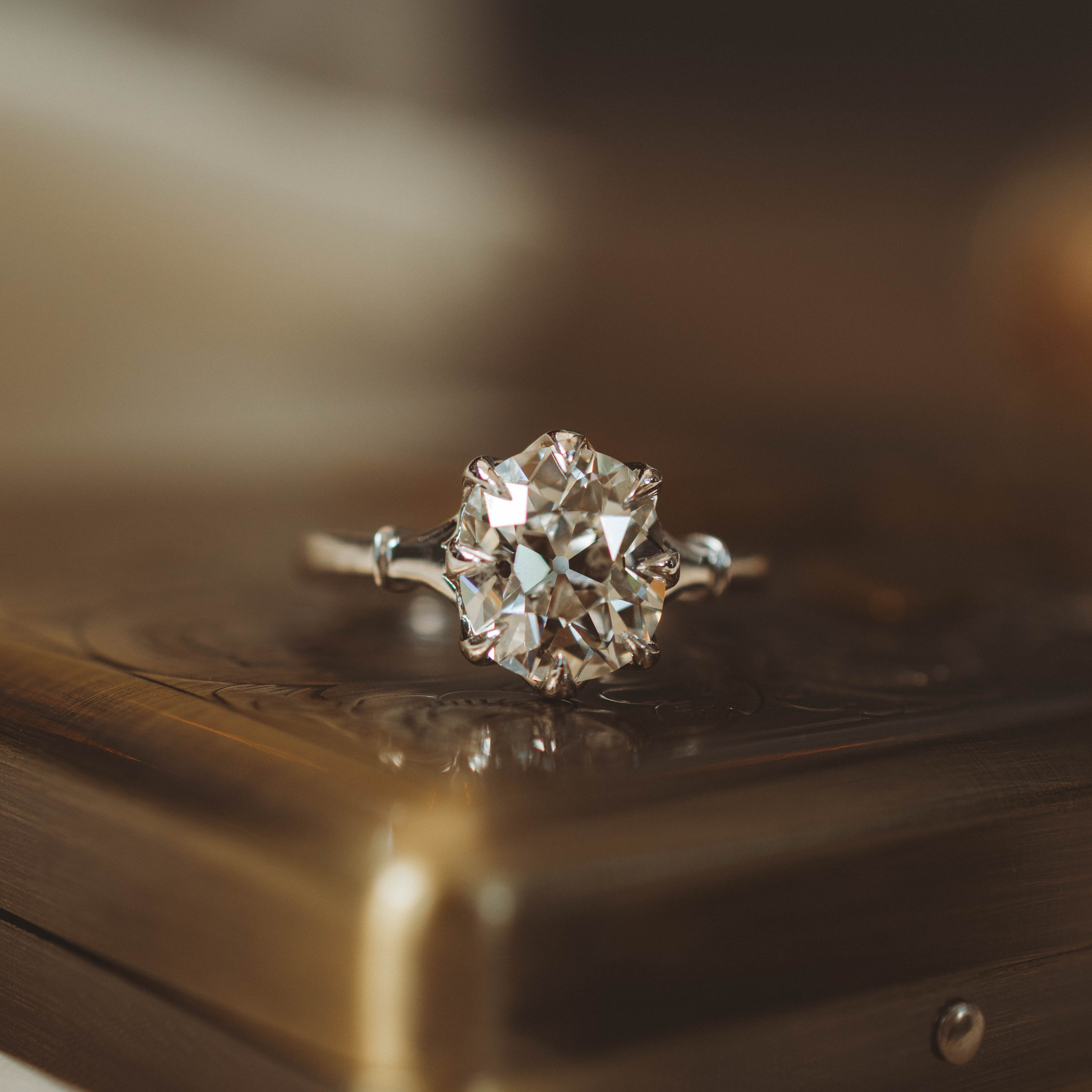 A close-up shot of a cushion old mine cut diamond engagement ring with a silver-toned band, resting on a polished bronze-colored metal surface.