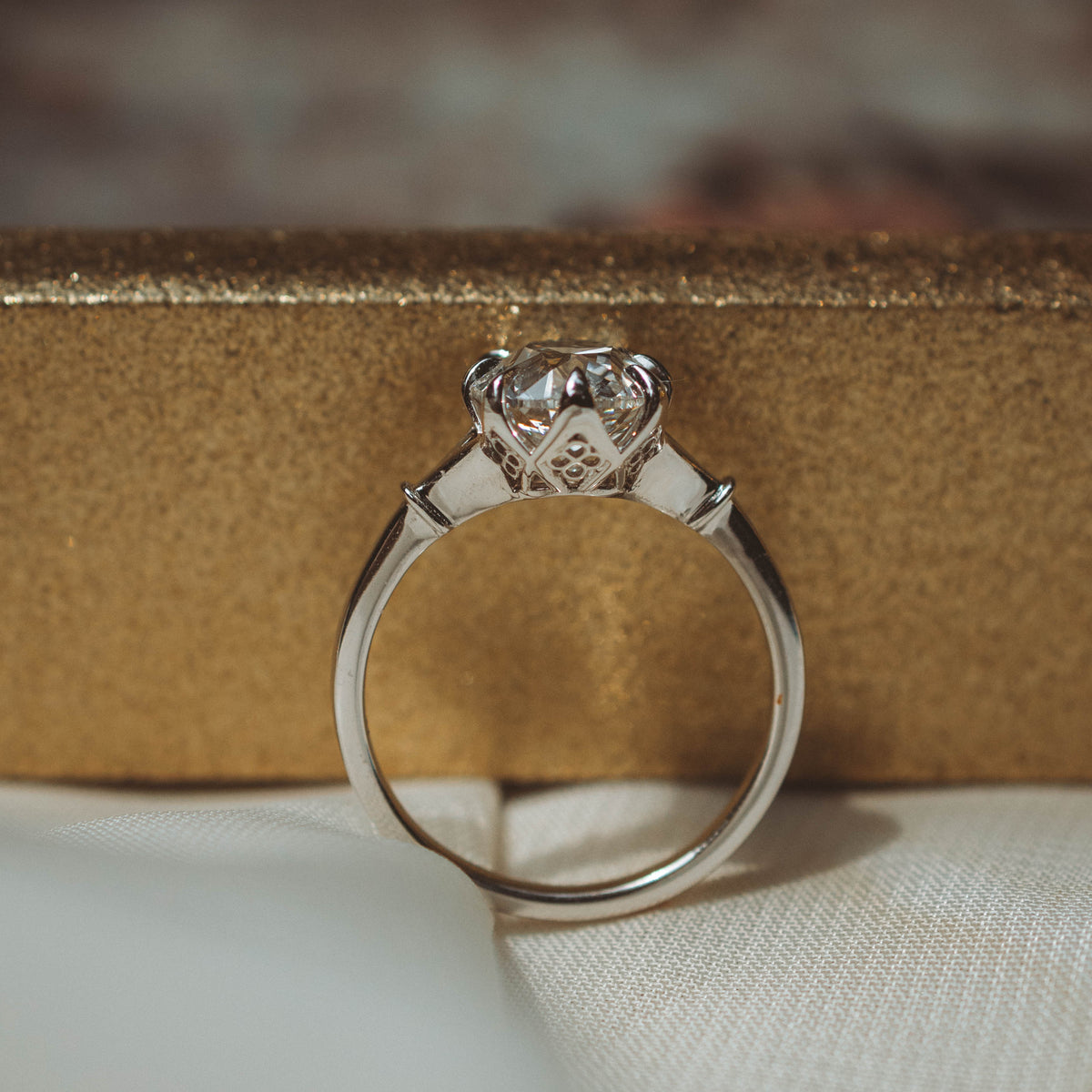 A white gold ring with an antique cut diamond in a prong setting, resting on a textured gold surface against a soft white fabric background.