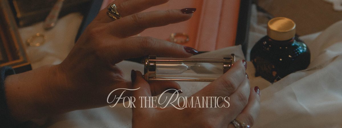 A close-up of a person's hands holding a small rectangular metallic object, with rings on the fingers and a dark bottle nearby on draped white fabric. The text 'For The Romantics' is overlaid across the center.