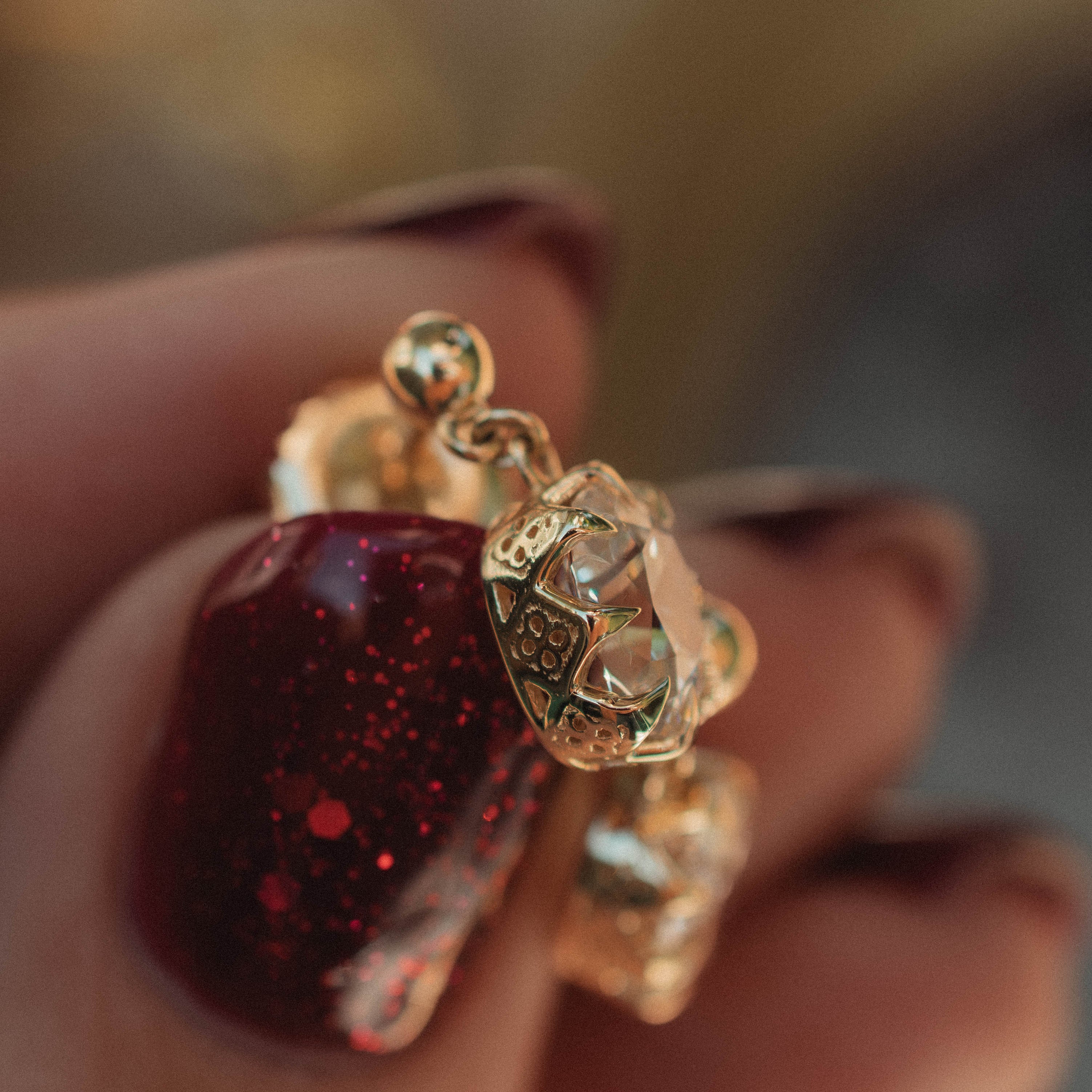 A close-up of a hand with dark red glitter nail polish holding a gold ring with a faceted clear gemstone.