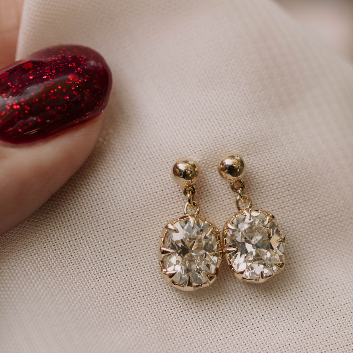 A pair of gold drop earrings with large clear round-cut stones, resting on a beige fabric surface next to a hand with glittery red nail polish.