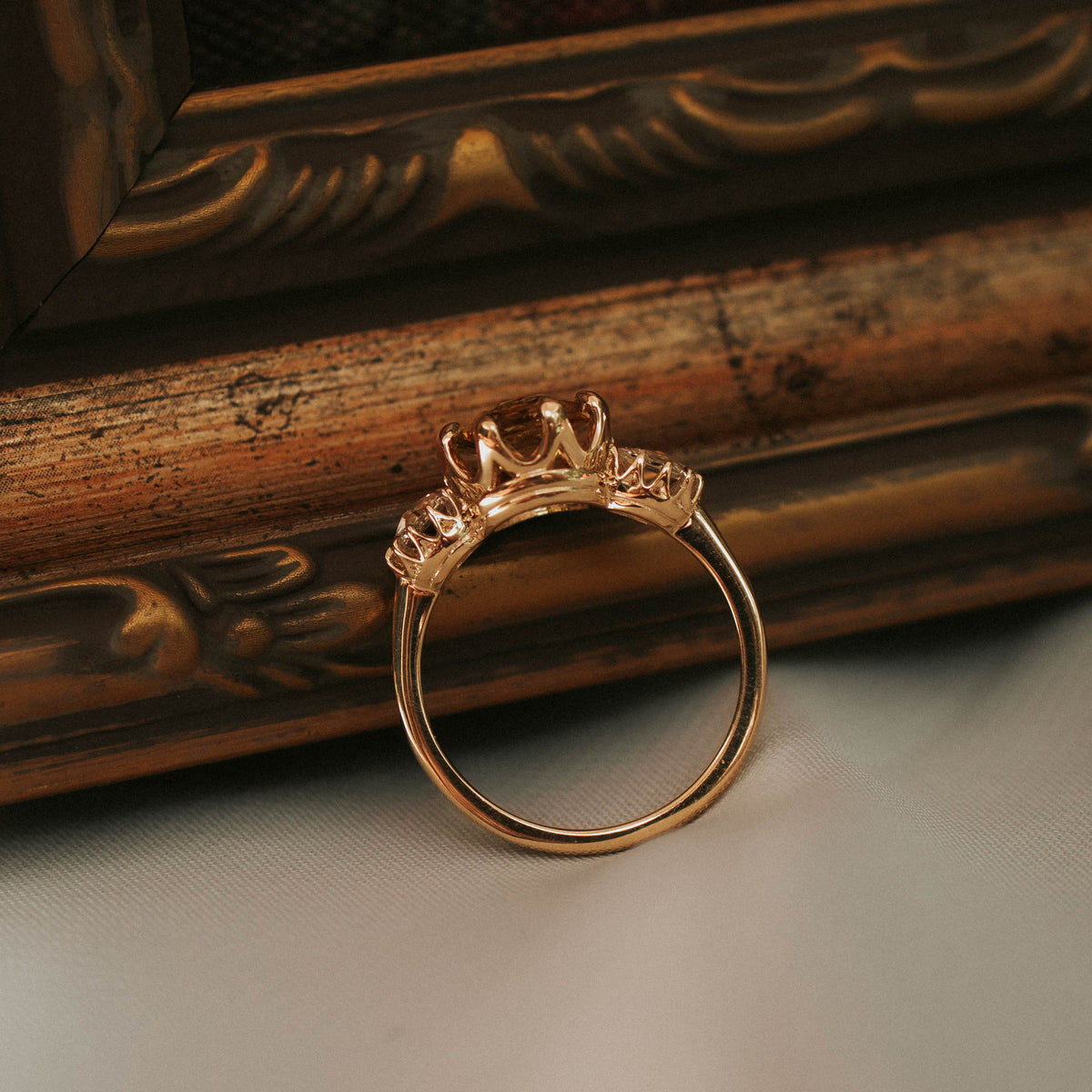 A gold ring with a crown-like top setting, photographed resting against an ornate vintage picture frame on a light surface.