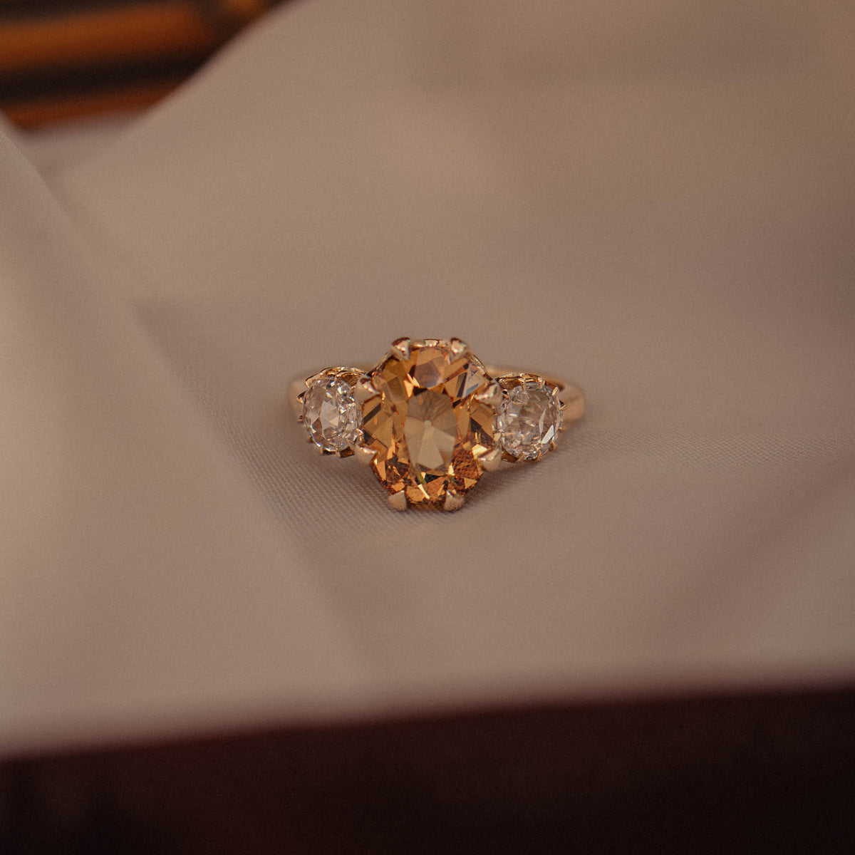 A yellow gold ring with a central oval orange gemstone and two small round rose cuts resting on a white surface with a dark wooden background.