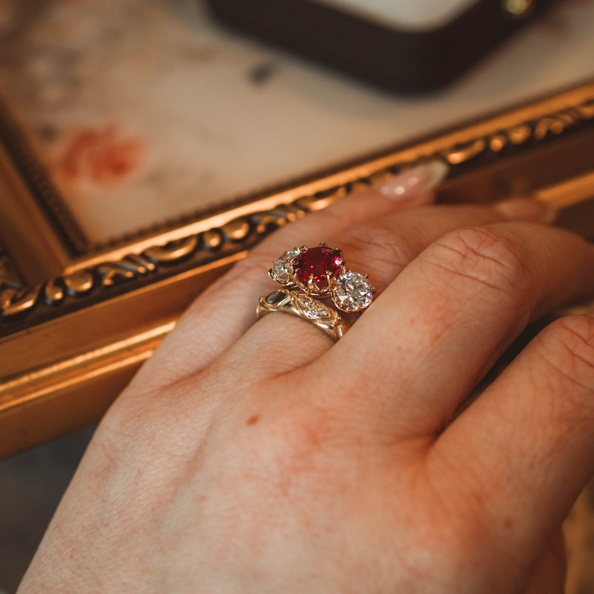 ruby and diamond three stone trilogy engagement ring worn on hand with wedding band wiht ring box and painting background.