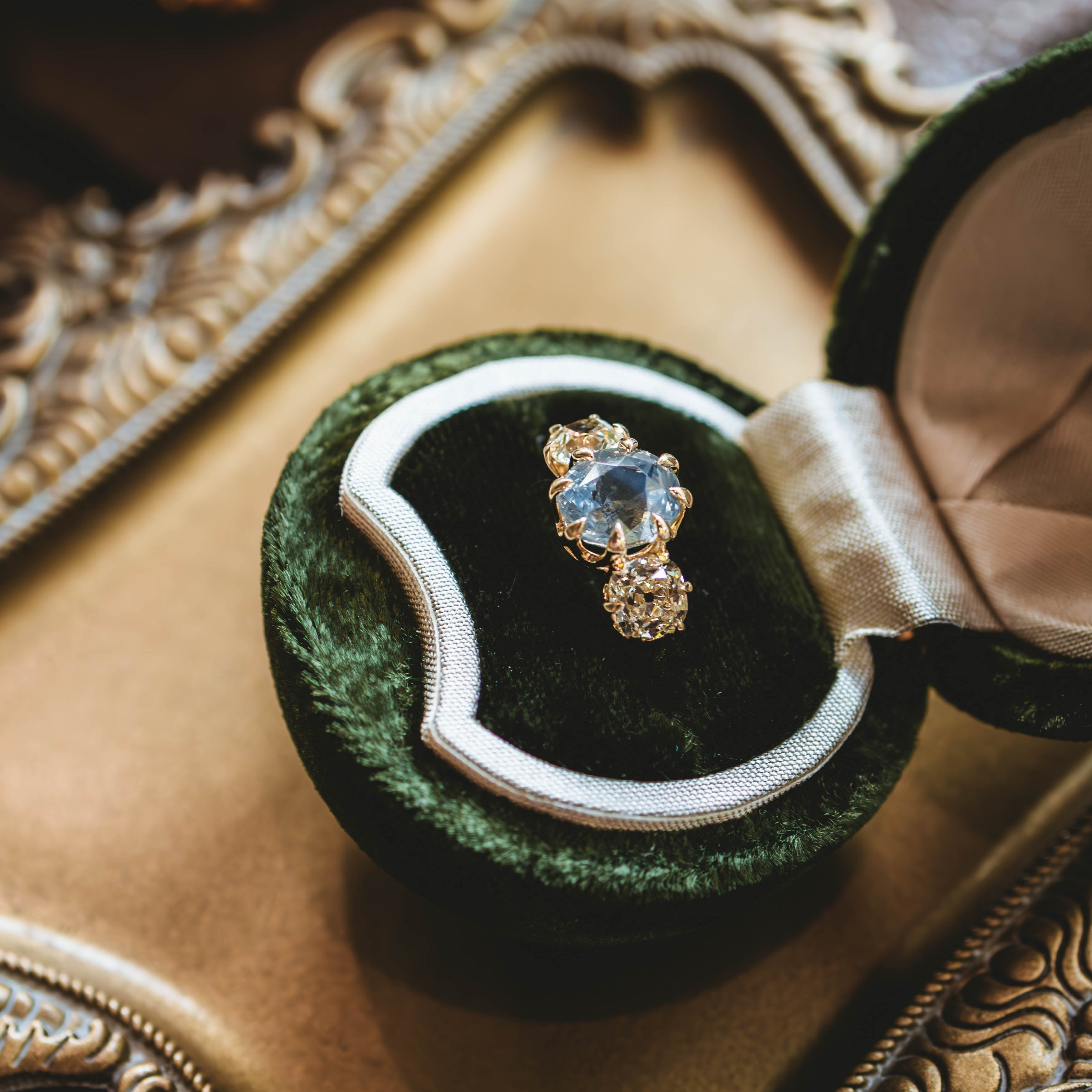 An open dark green velvet ring box holding a gold ring with a light blue oval gemstone, placed on an ornate gold tray.