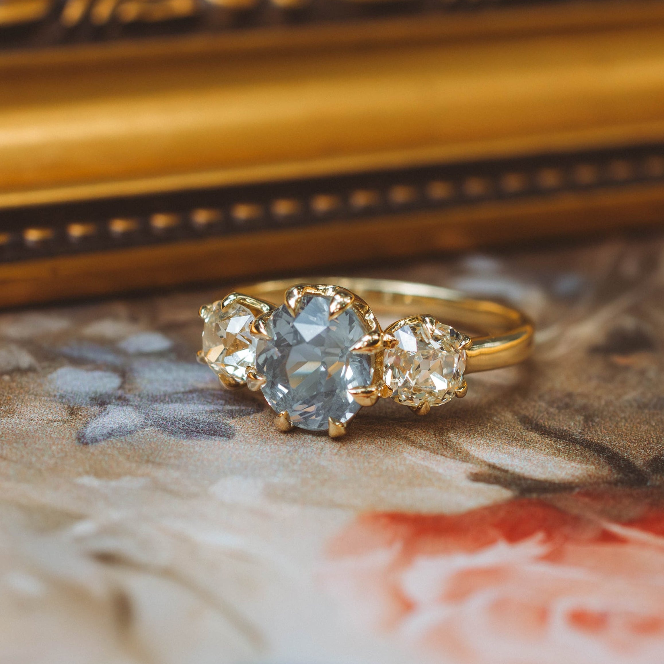 A gold three-stone ring featuring a central round light blue gemstone and two small clear accent stones, resting on a floral surface with a blurred ornate gold frame in the background.