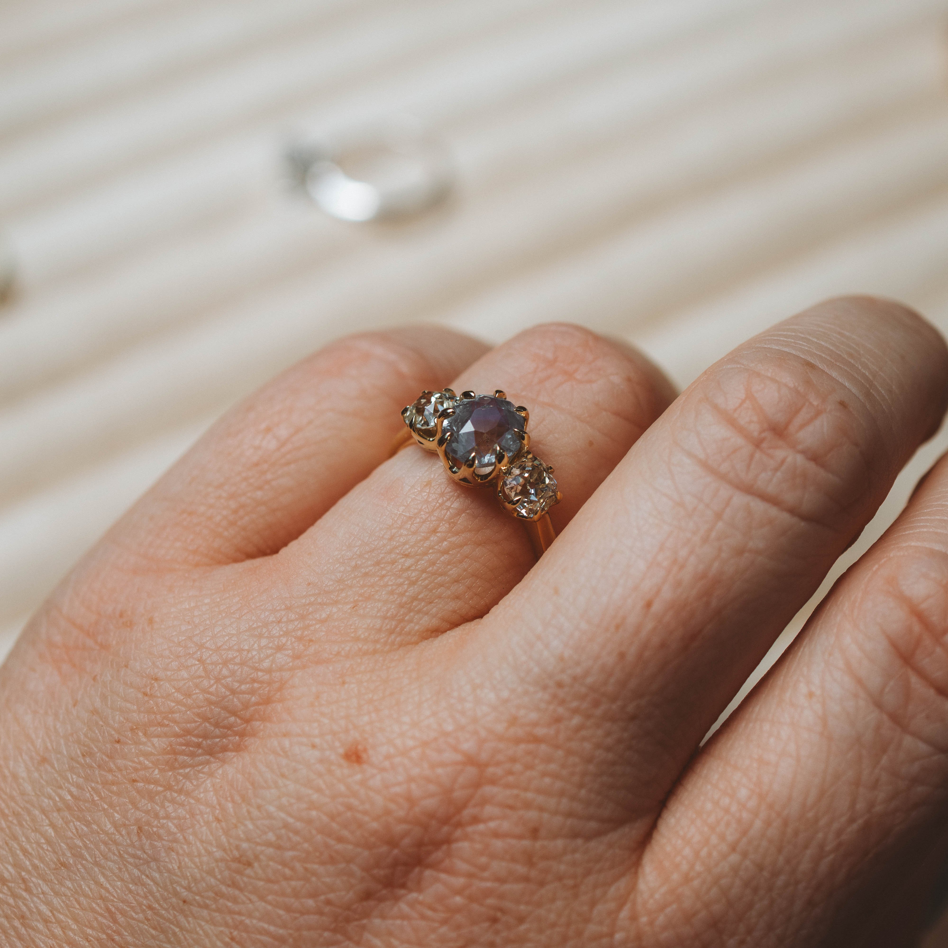 A close-up of a hand wearing a vintage-style gold ring with a central oval blue gemstone and small side stones, set against a softly blurred cream-colored background.