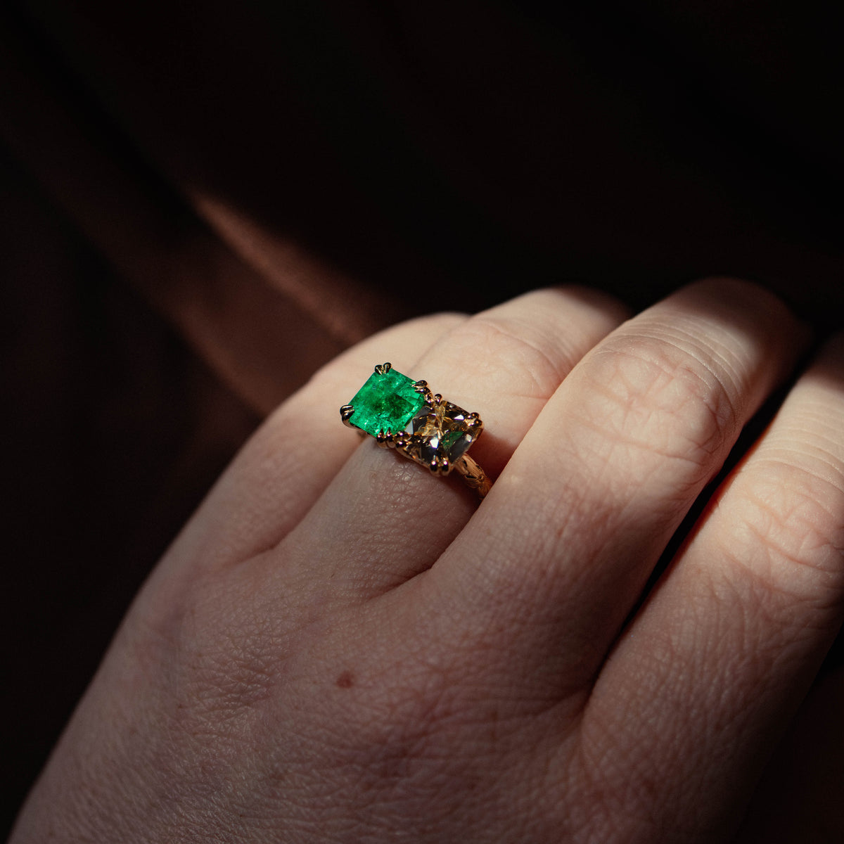 A close-up of a hand wearing a gold ring with a square-cut green emerald gemstone and cushion warm diamond, set against a dark, shadowed background.