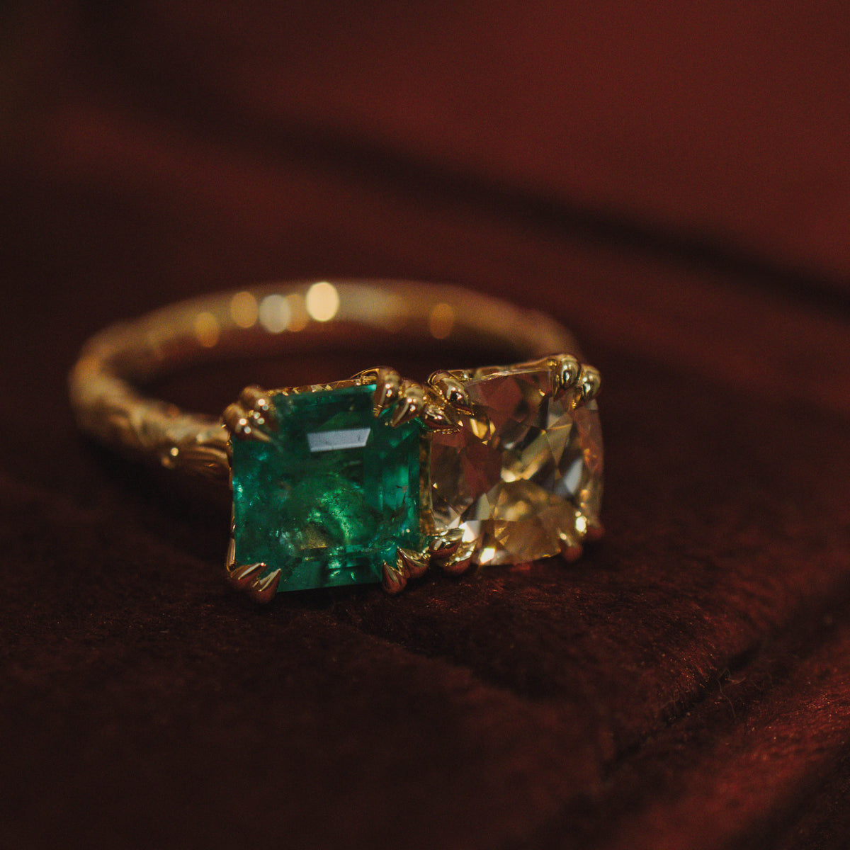 A gold ring featuring a square-cut green emerald gemstone and a cushion-cut pale yellow diamond, resting on a dark brown velvet surface.