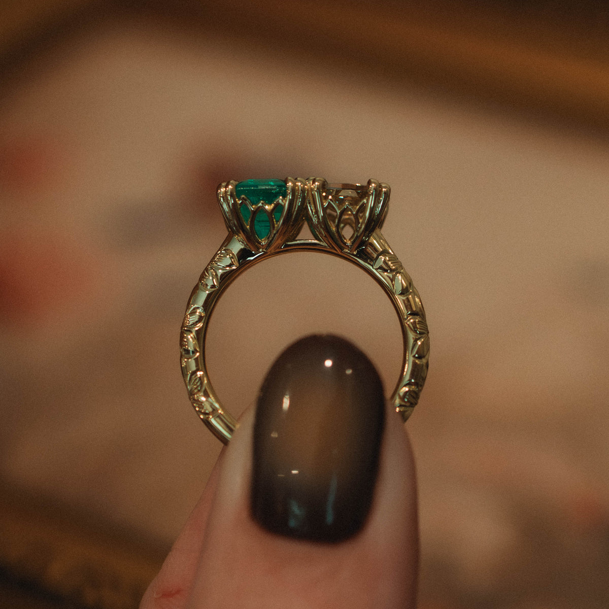 A close-up of a gold ring with a green emerald and warm diamond in ornate arch baskets held between fingers with dark smoky nail polish against a blurred warm-toned background.