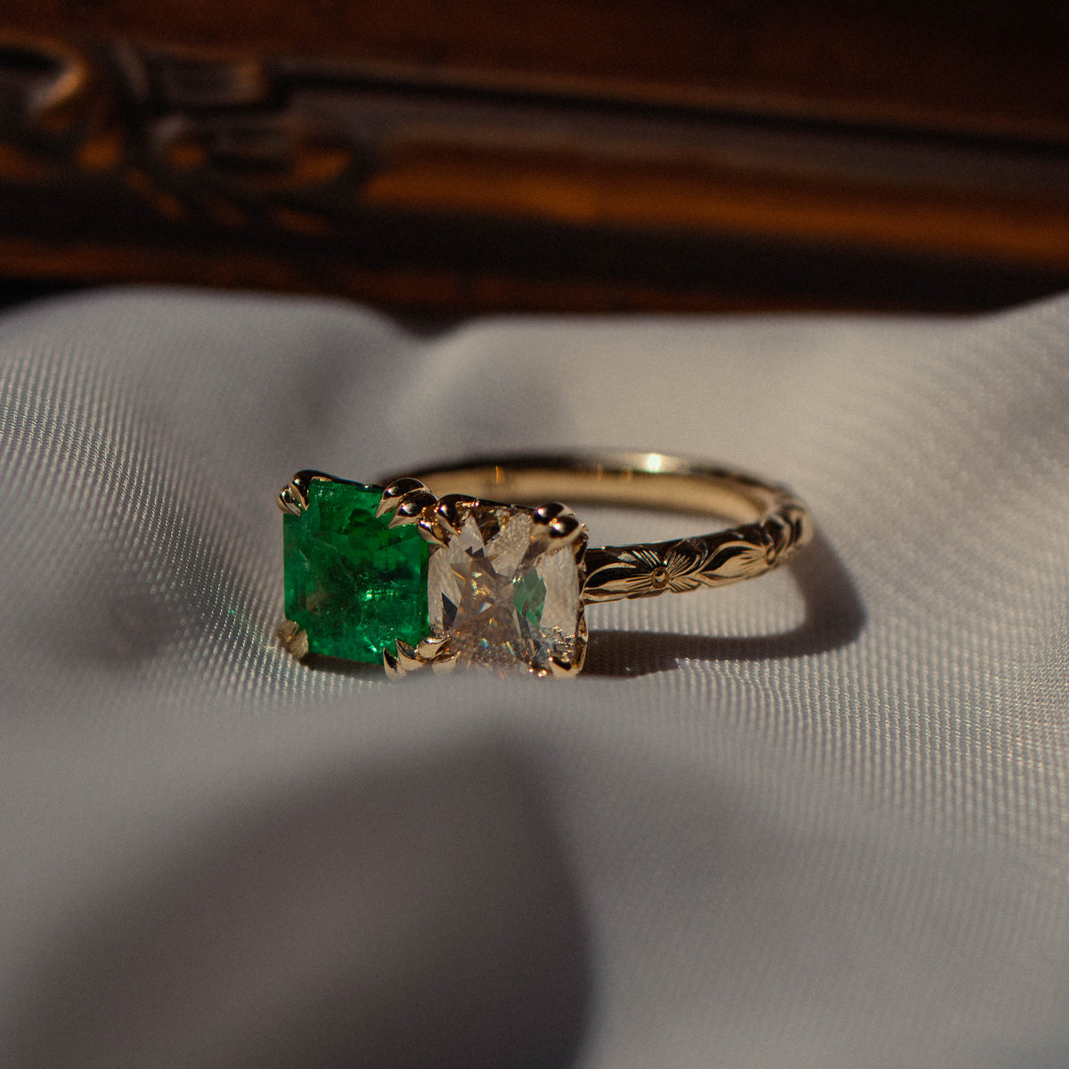 A gold ring featuring a square-cut green emerald gemstone and a warm cushion diamond resting on a white textured fabric surface with a dark wooden background.