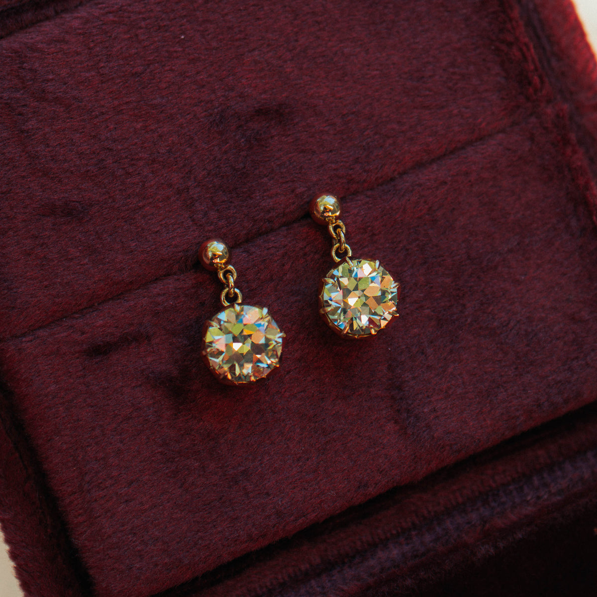 A pair of gold drop earrings with round, faceted diamonds resting on a dark burgundy velvet jewelry box.