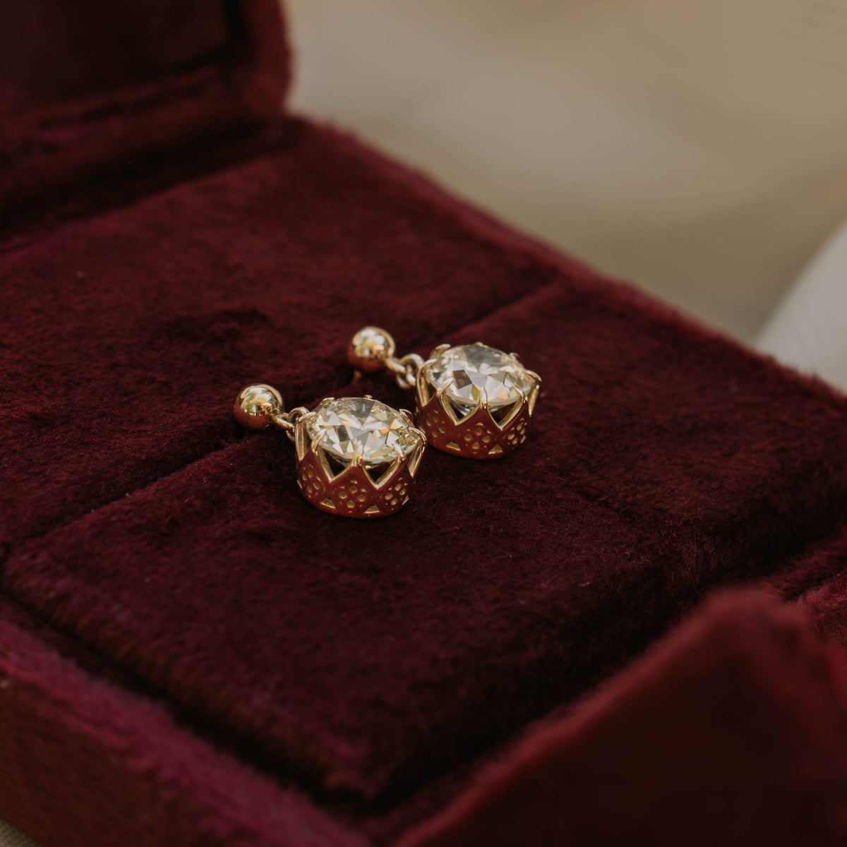 A pair of gold-toned statement earrings with warm faceted diamonds and gothic motif basket settings sitting in red velvet jewelry box