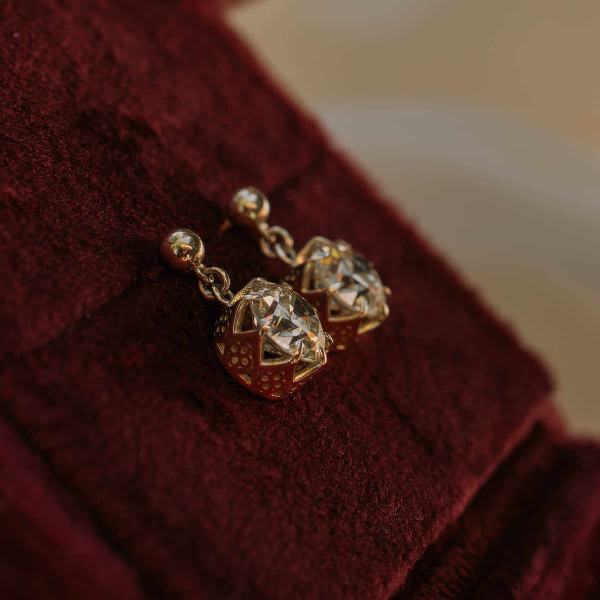 A pair of gold geometric drop earrings with clear faceted stones, resting on a deep burgundy velvet jewelry box.