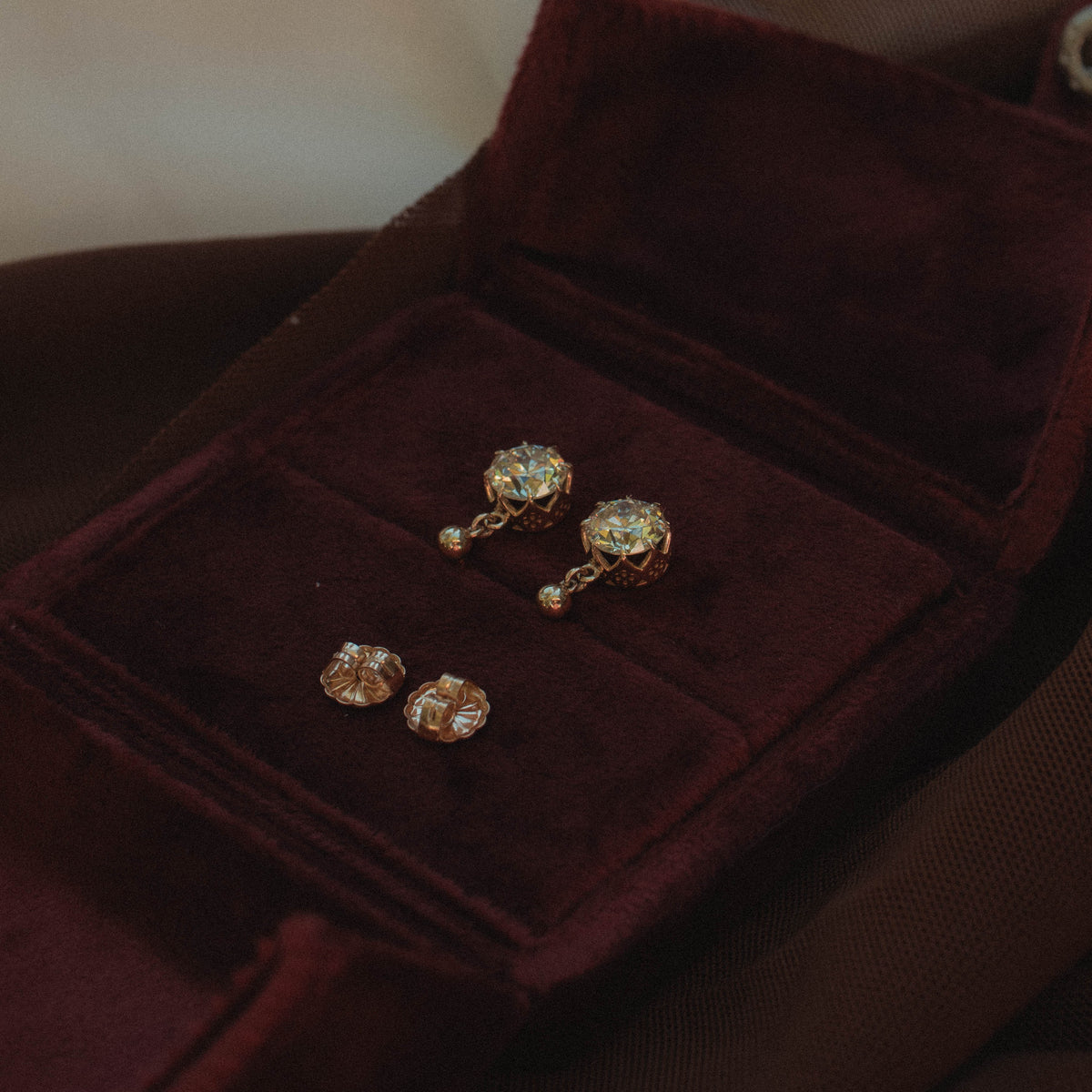 A pair of gold-toned stud earrings with clear gemstones displayed on an open dark burgundy velvet jewelry box, set against a soft beige fabric background.
