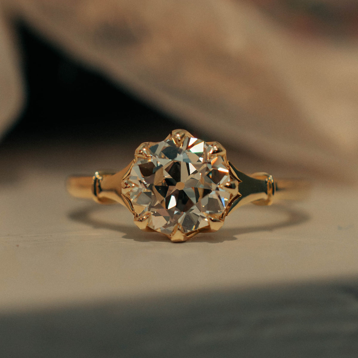 A vintage-style gold ring with a large round-cut old mine diamond in a floral cluster setting, resting on a textured surface with a soft-focus background.