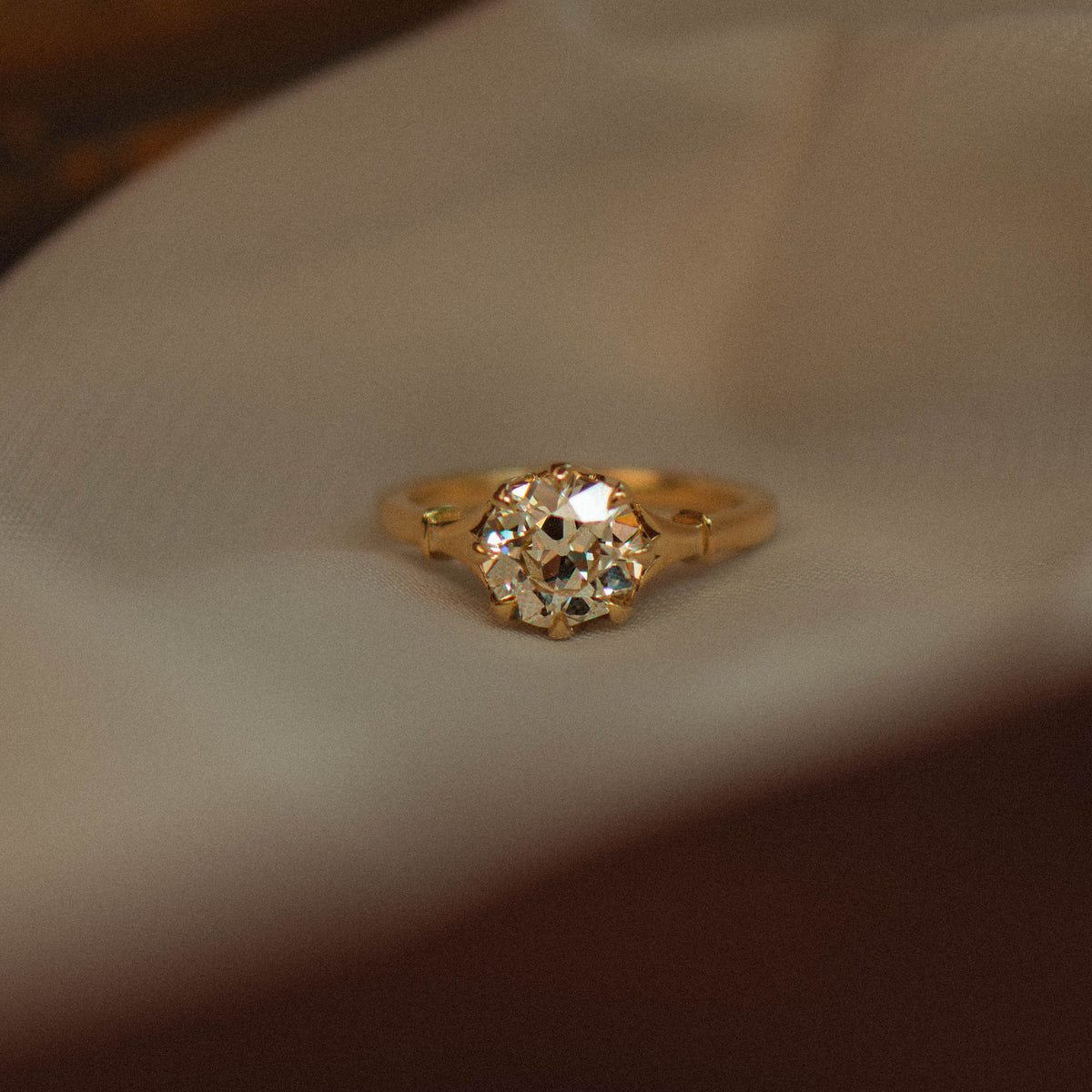 A gold ring with a round old mine diamond in a prong setting, resting on a white textured fabric surface with a dark, blurred background.