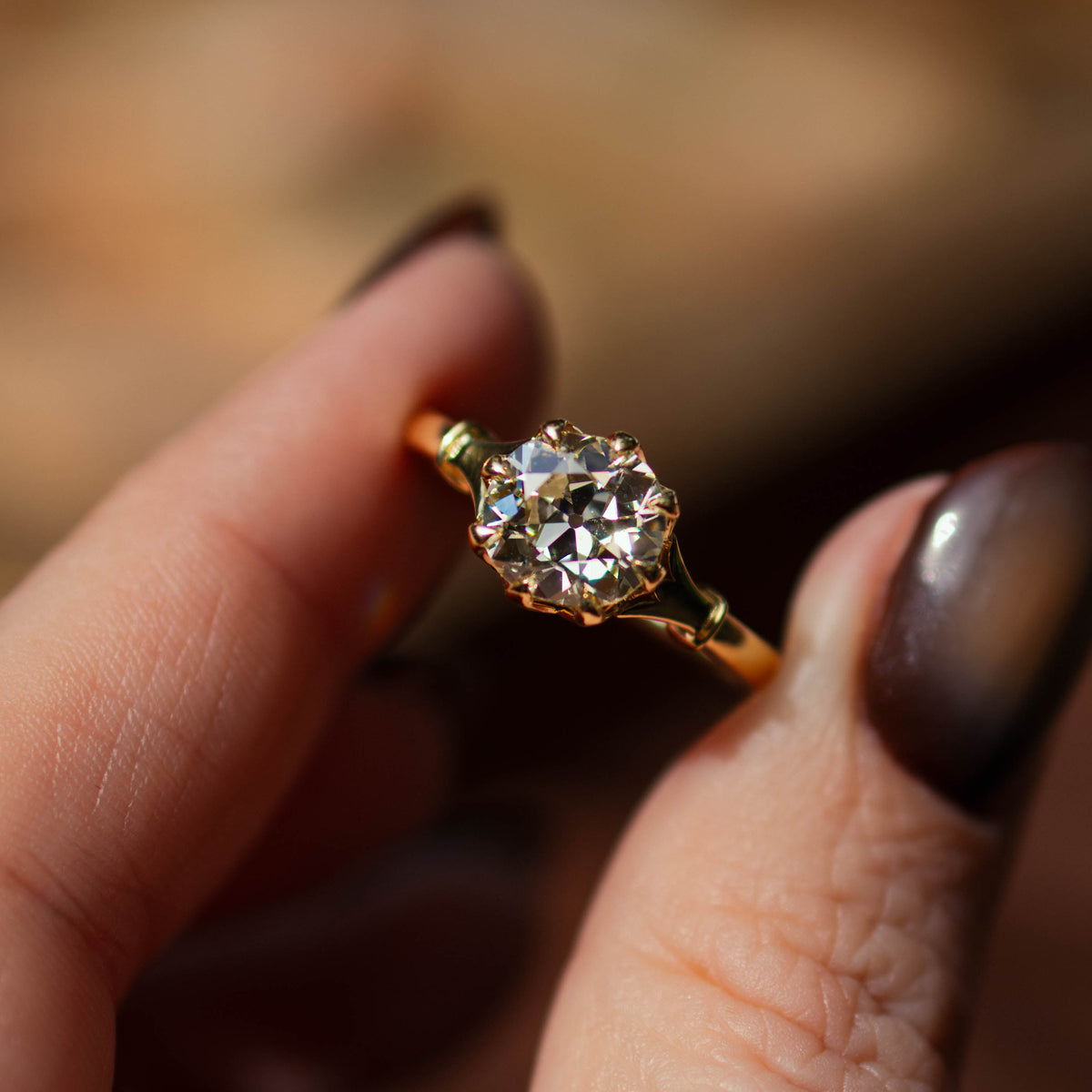 A close-up of a hand with dark metallic nail polish holding a gold ring with a round-cut old mine diamond gainst a warm, blurred background.