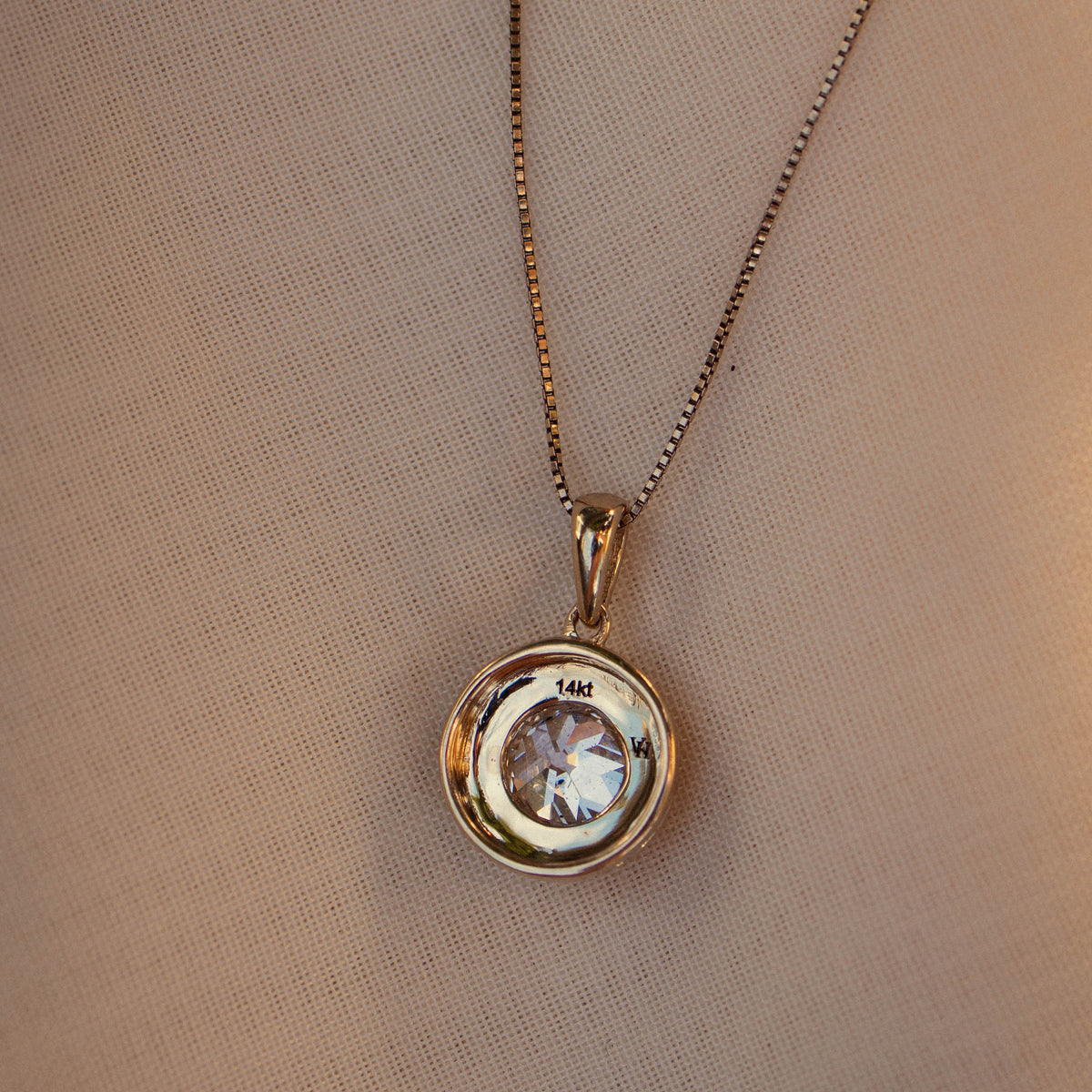 A gold pendant necklace displayed on a textured beige fabric background.
