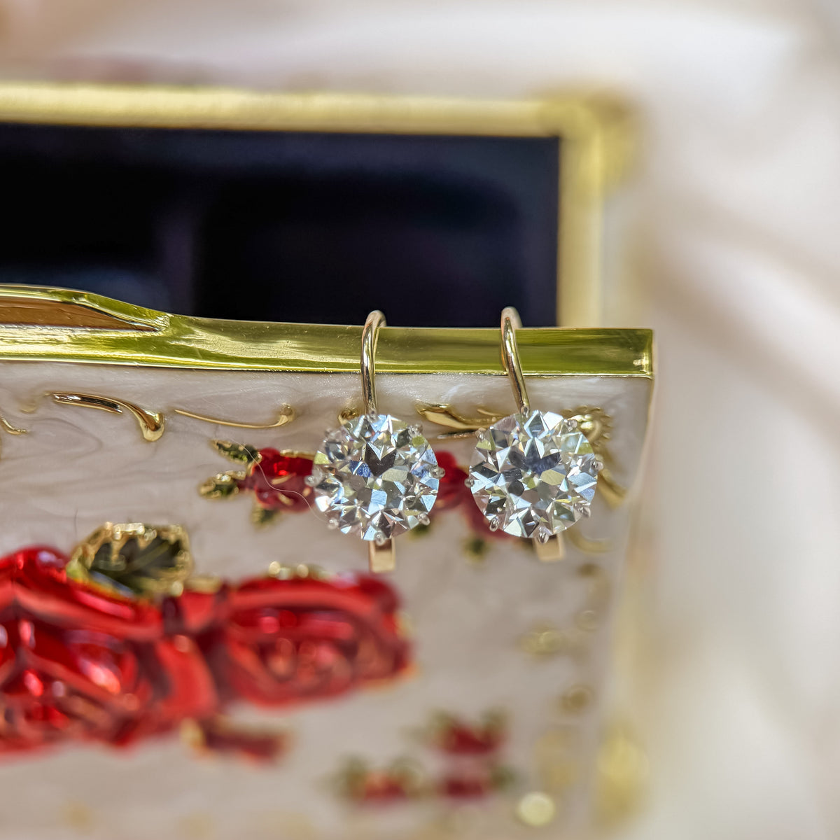 A pair of gold hoop earrings with round old european cut diamonds, resting on a decorative white and gold box with red rose details, set against a soft white fabric background.