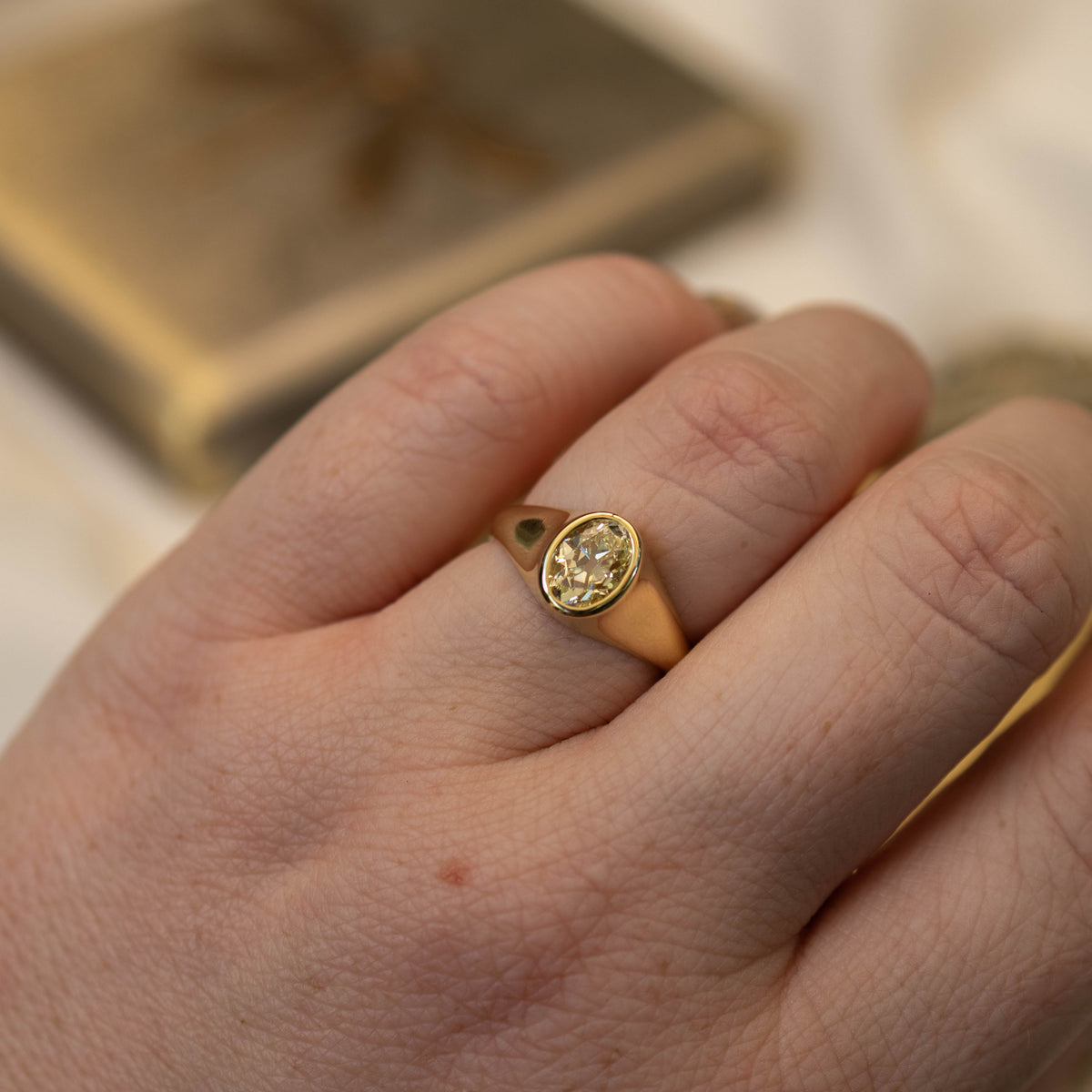 A close-up of a person's hand wearing a gold signet ring with an oval yellow diamond gemstone, set against a soft, out-of-focus background.
