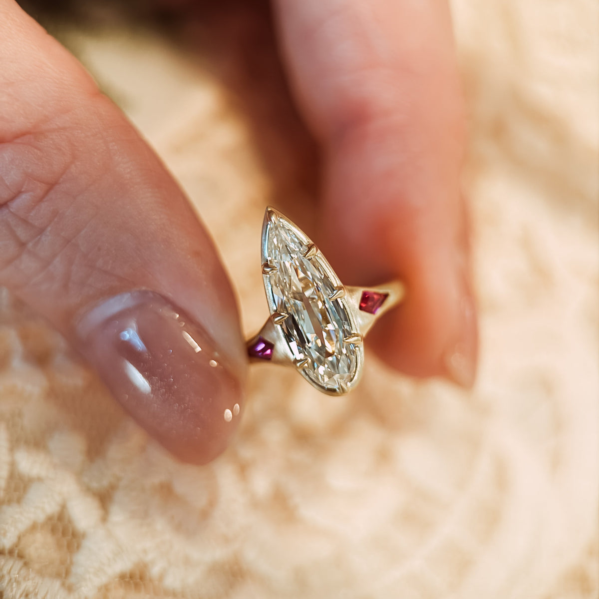A close-up of a hand wearing a gold ring with a pear diamond and small red accent stones, resting on a beige lace surface.