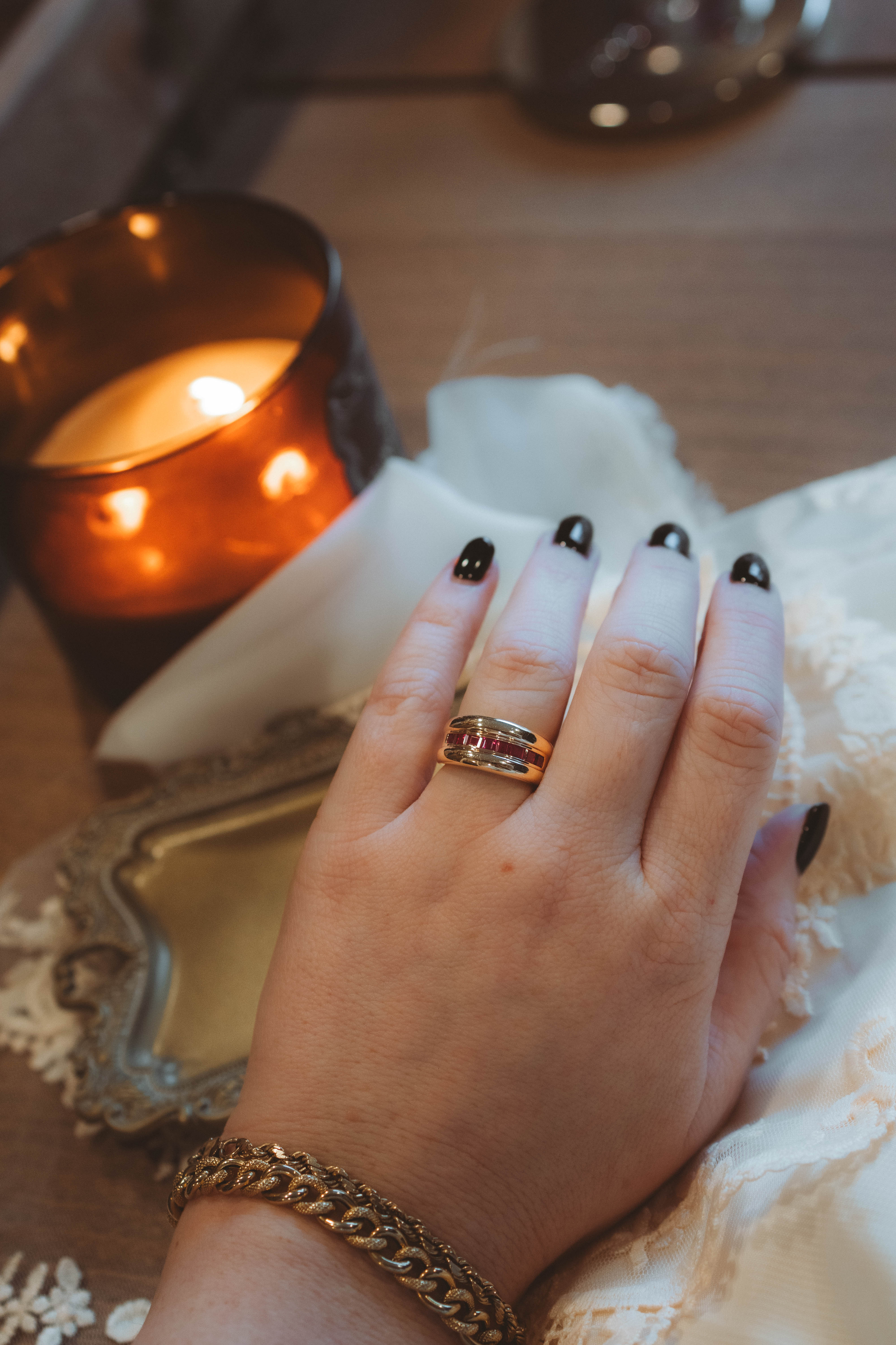 Hand with a chunky gold and ruby ring on a decorative tray with a lit candle in the background
