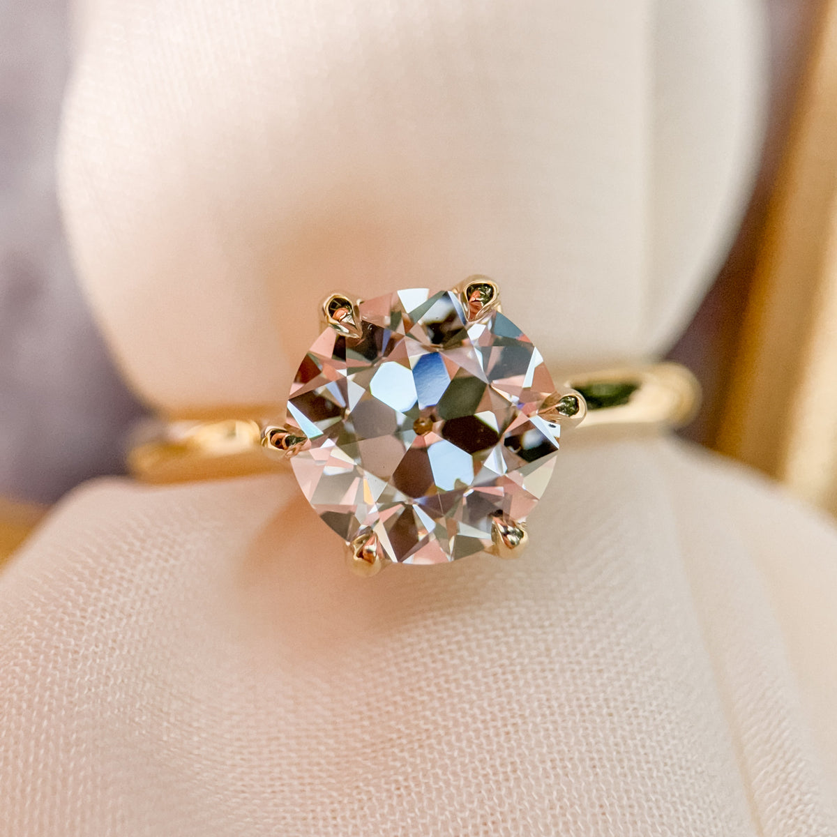 A yellow gold solitaire ring with a large round old european diamond displayed on a soft cream-colored fabric ring holder.