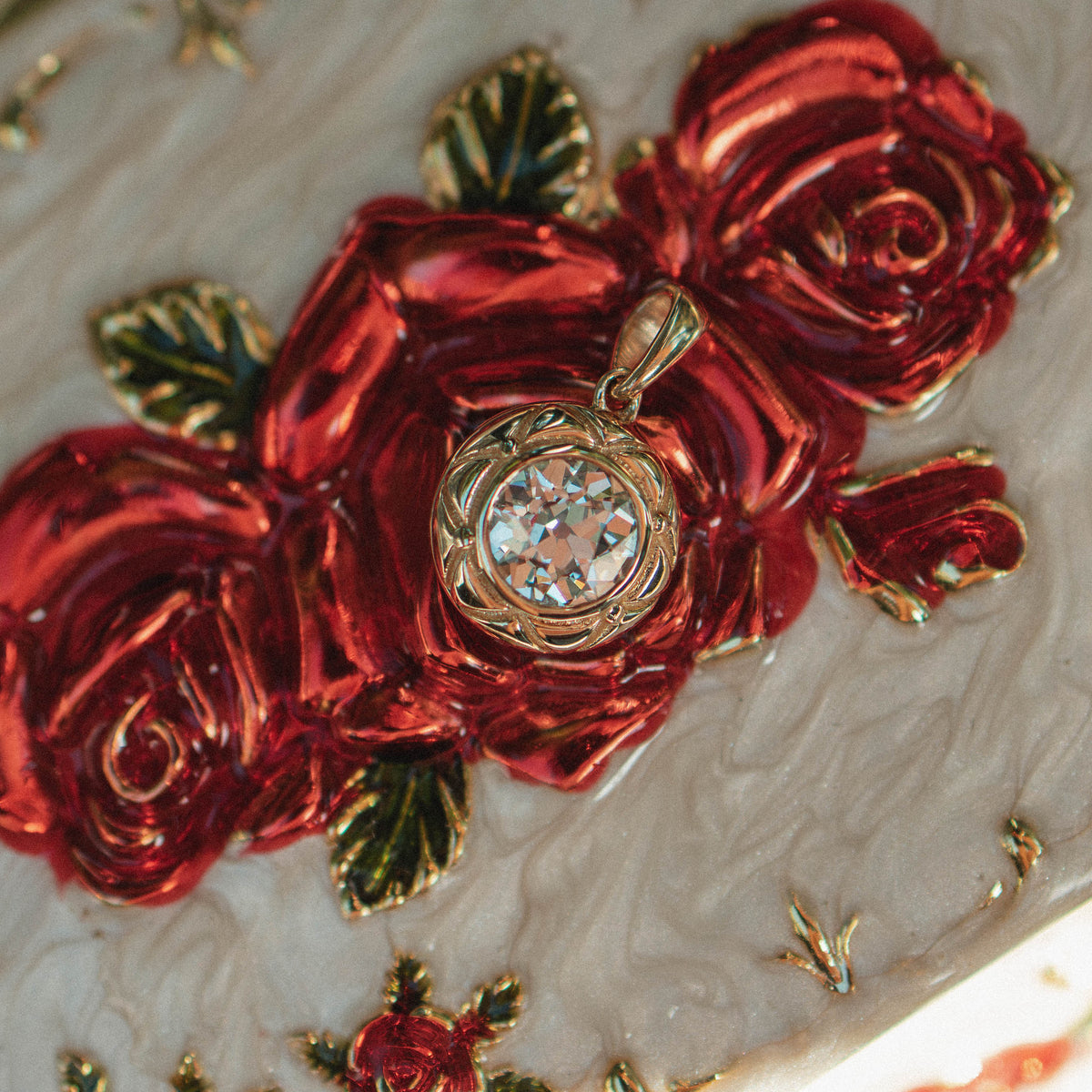 A close-up of an ornate white embossed case decorated with large glossy red roses, green leaves, and gold floral detailing, featuring a central round gold pendant with a round old euroean cut diamond.