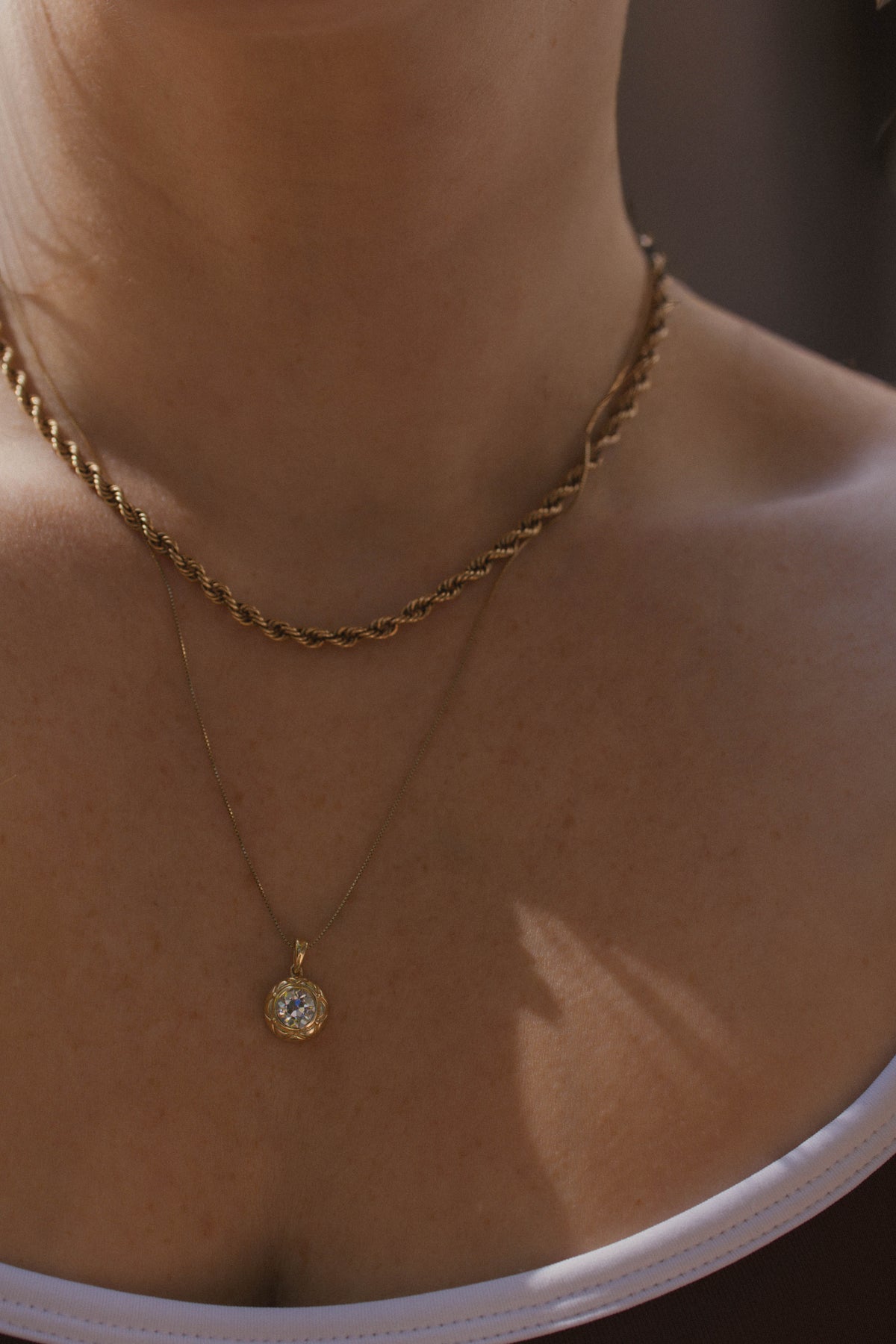 A close-up of a person's neck and upper chest wearing layered gold necklaces, including a twisted chain and a delicate chain with a small round pendant.