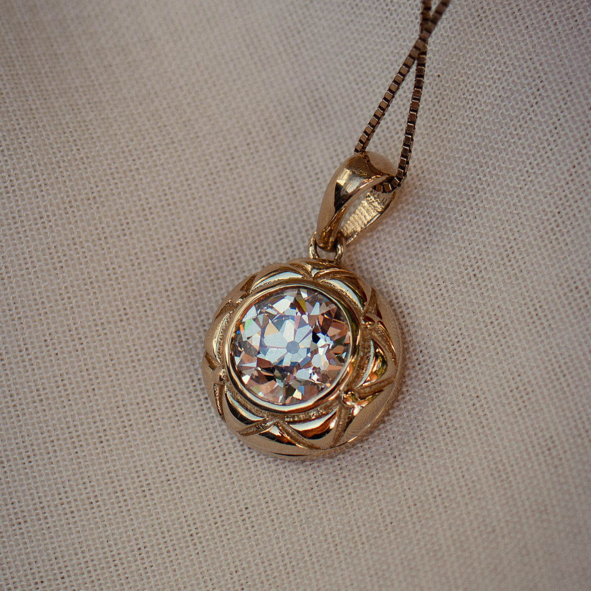 A gold pendant necklace featuring a large, round old european diamond surrounded by a textured, swirling gold setting, resting on a neutral beige fabric background.