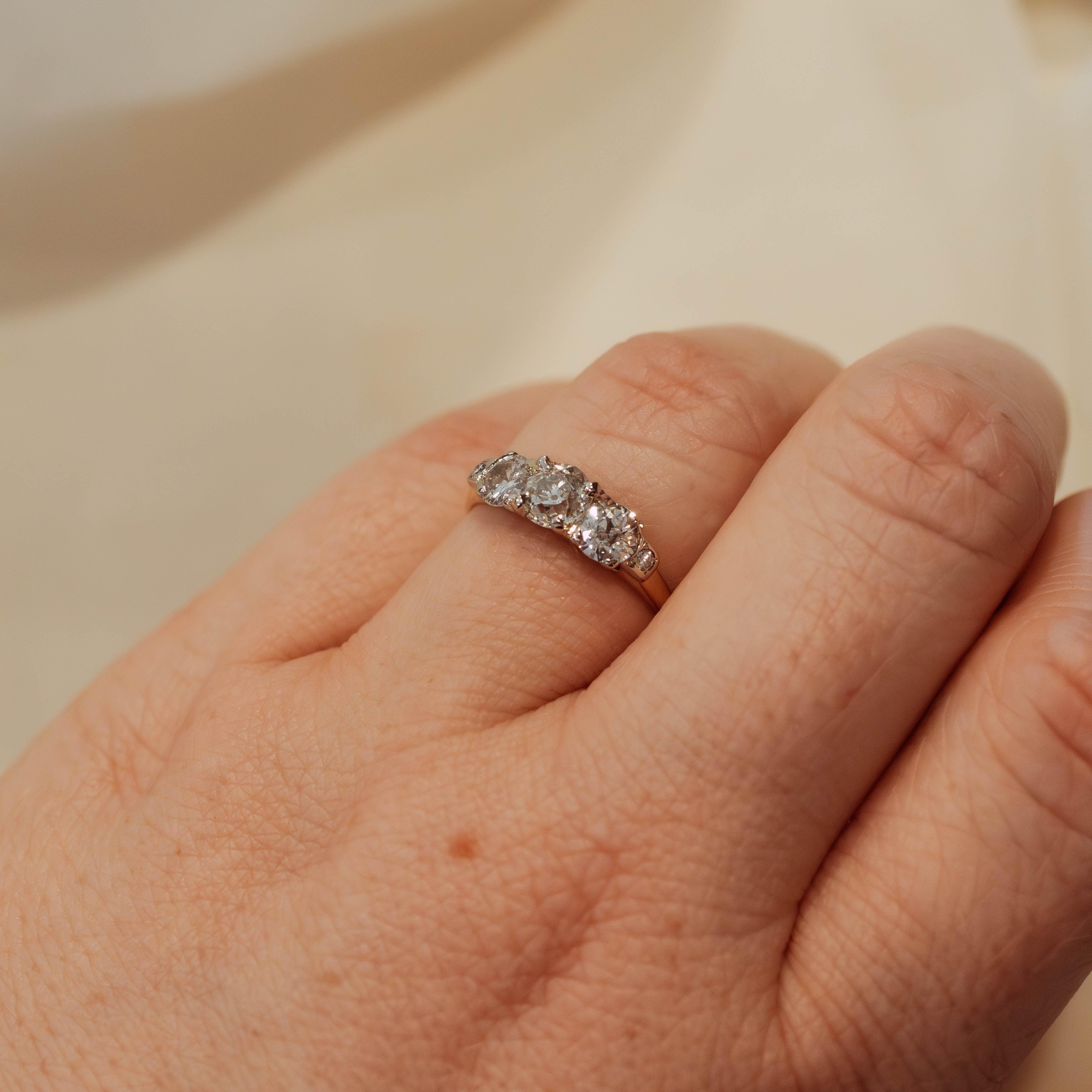 A close-up of a hand wearing a gold three-stone diamond ring against a soft cream-colored fabric background.
