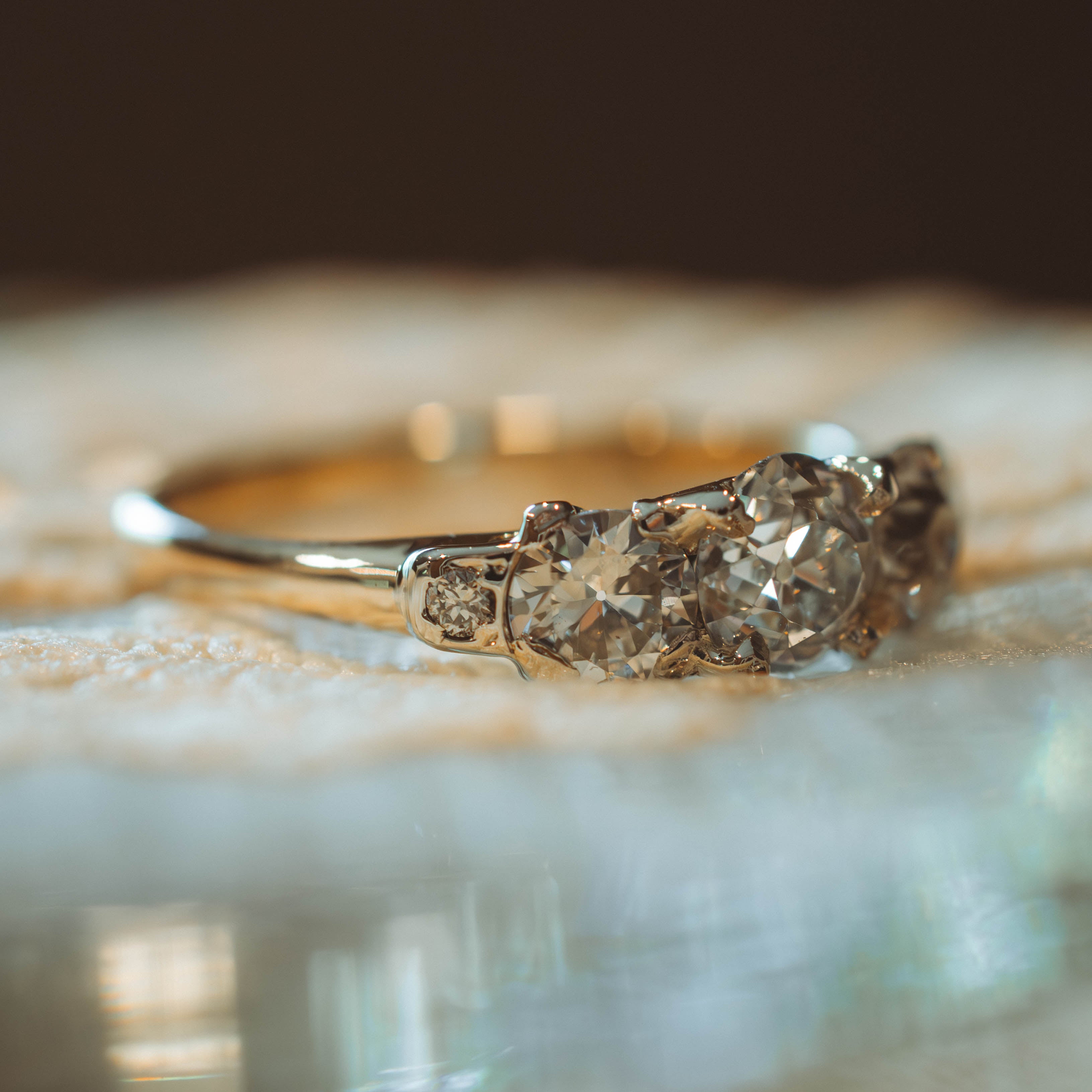 A close-up of a vintage-style three-stone diamond ring with a gold band, resting on a textured white surface with a soft-focus background.