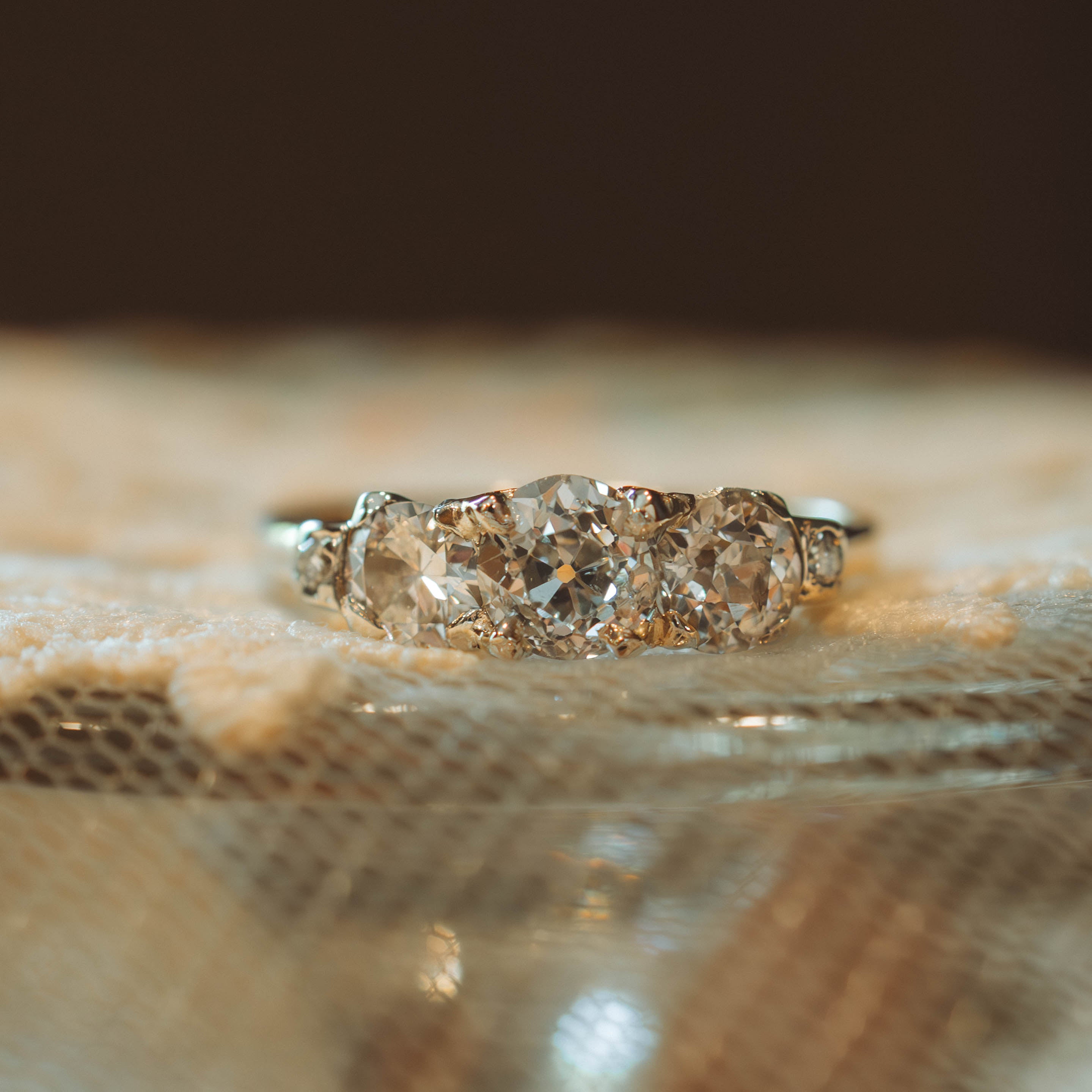 A three-stone diamond ring with a silver & gold toned band, resting on a textured beige fabric surface with a dark, blurred background.