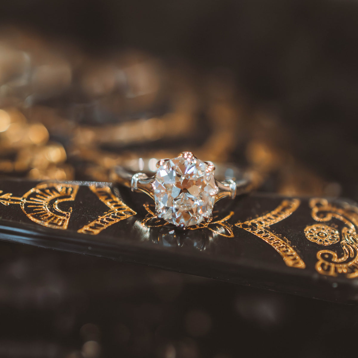 a cushion old mine cut diamond engagement ring set in a silver-toned band, resting on a glossy black surface with gold detailing.