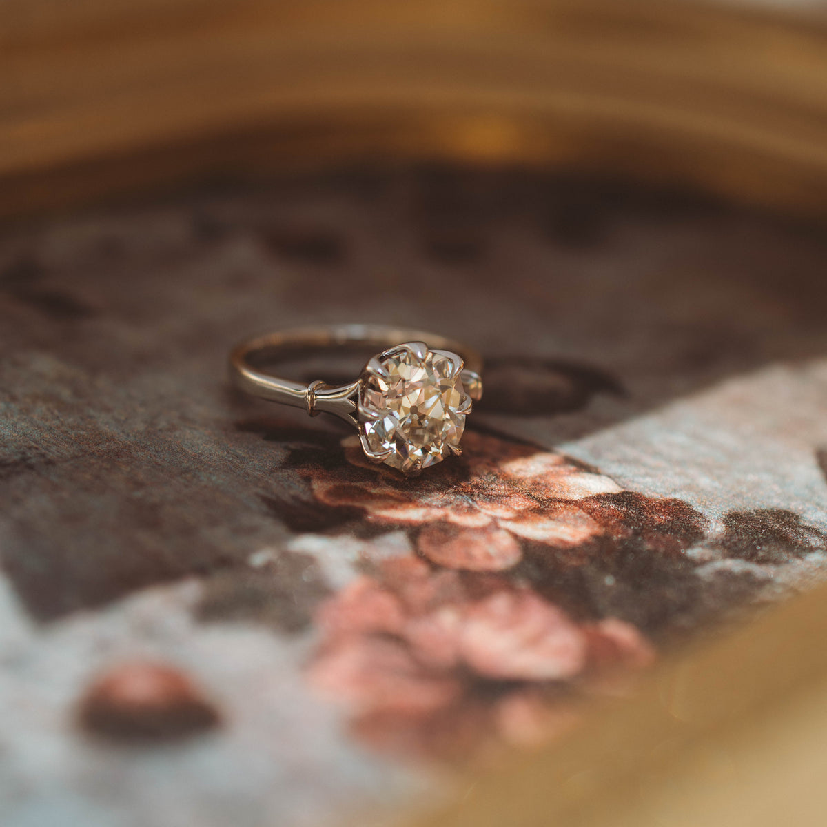 A vintage-style diamond engagement ring with a silver-toned band and a cushion old mine diamond , resting on a textured, floral-patterned surface.