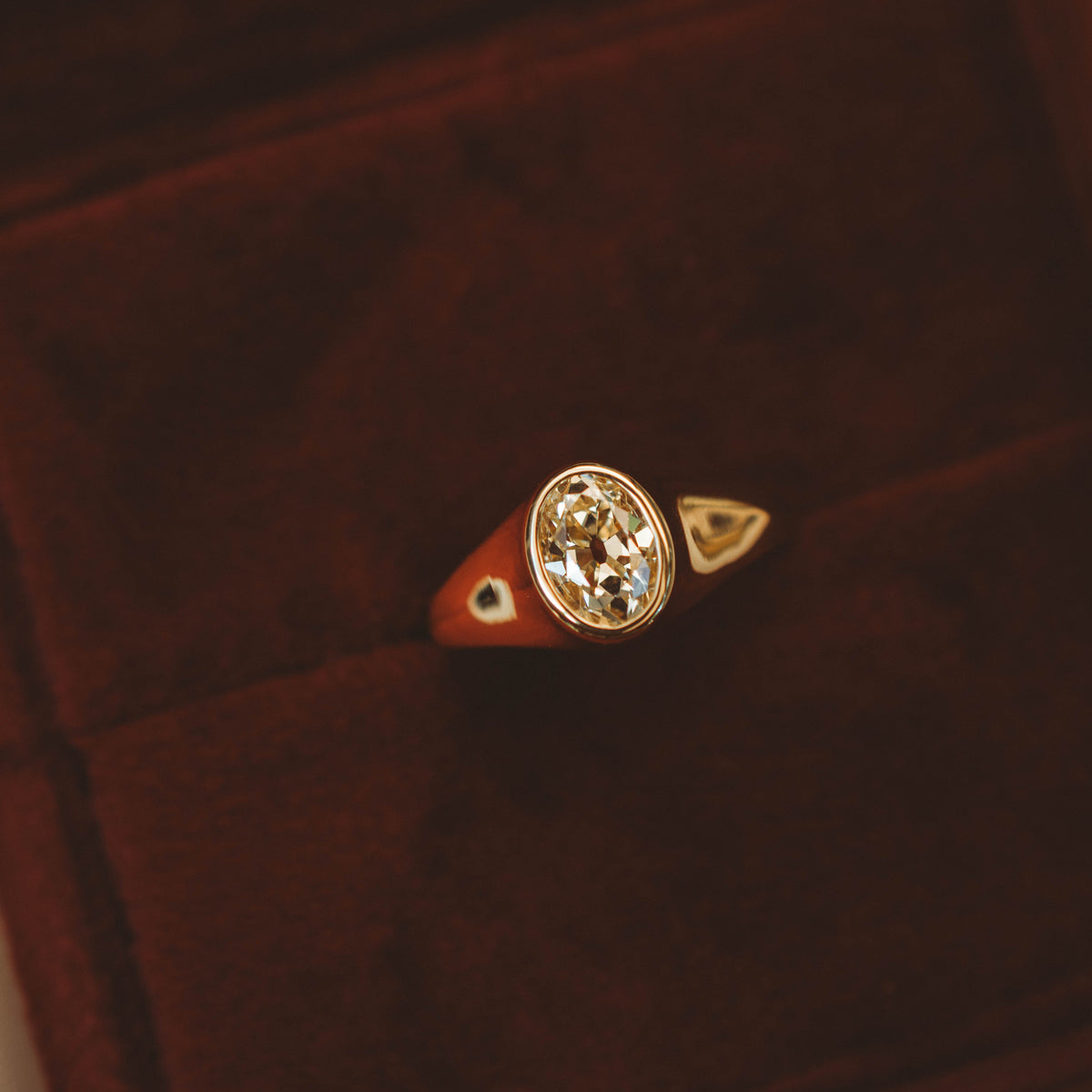 A gold signet-style ring with an oval diamond displayed in a dark brown velvet ring box.