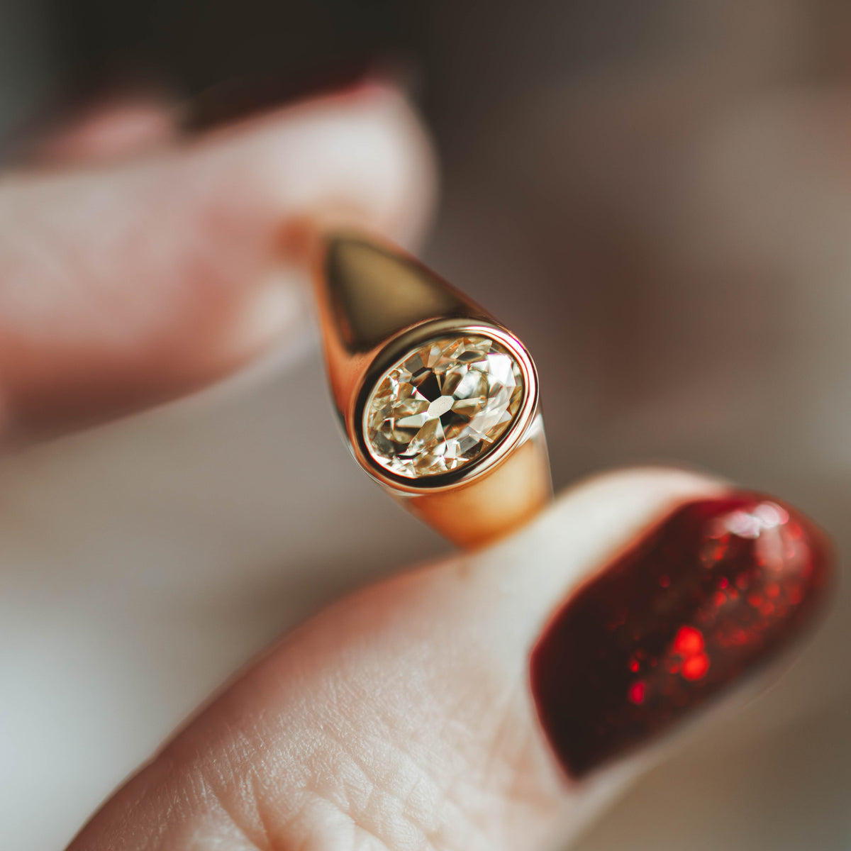 A close-up of a gold signet ring with an oval antique cut yellow diamond held between fingers with dark red glittery nail polish against a soft, blurred background.