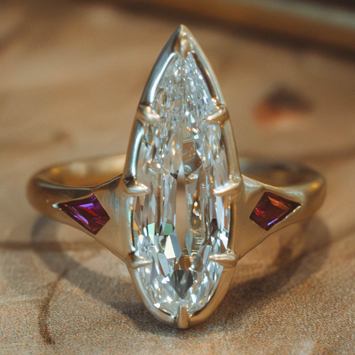 A gold ring featuring a large elongated pear cut diamonds in a prong setting, with two small red diamond-shaped side stones, photographed close-up on a textured brown surface.