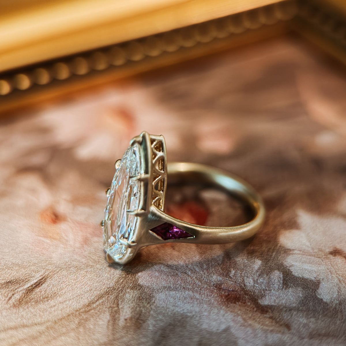 An antique-style gold ring with a pear diamond  and a small red gemstone accent, resting on a vintage floral print surface.
