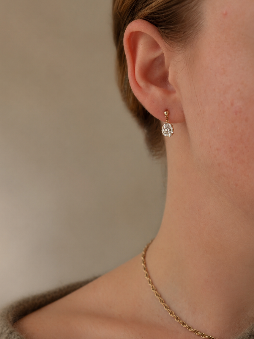 A close-up lifestyle shot of a model wearing a delicate gold chain necklace and a small gold hoop earring with a clear, faceted circular drop pendant. The model is wearing a textured taupe knit sweater against a soft, warm beige background.