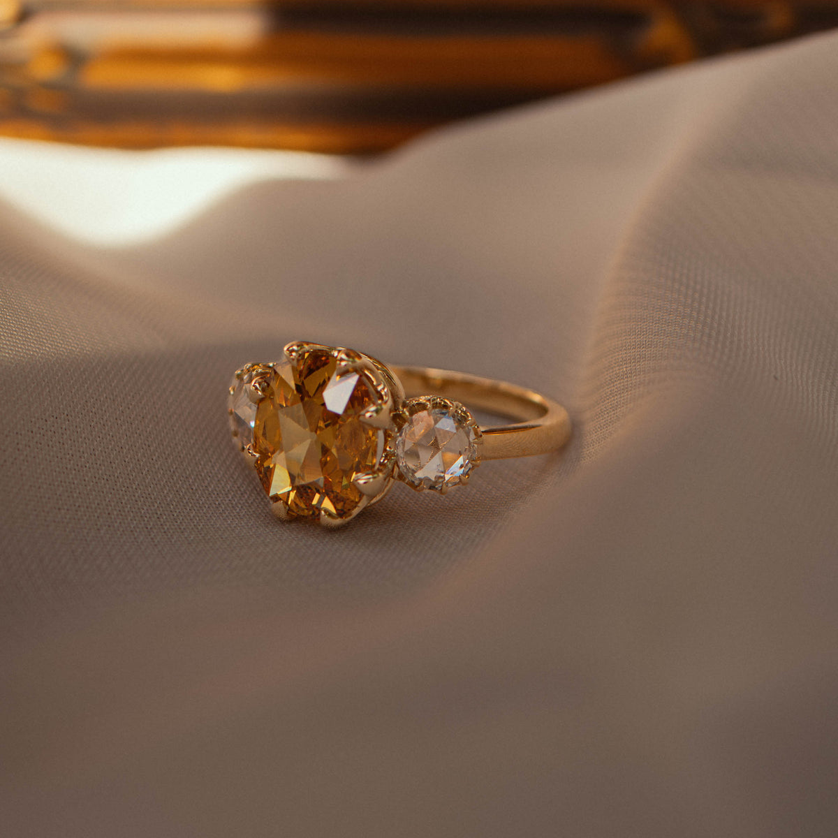 A gold ring featuring a large oval-cut yellow diamond and two smaller round rose cut diamonds, resting on draped light beige fabric with a warm, blurred golden background.