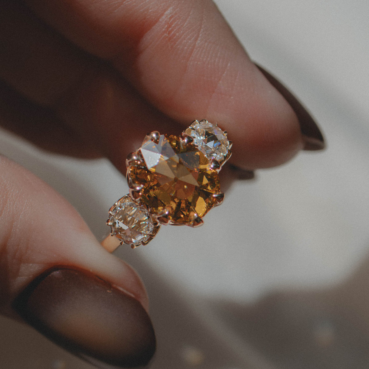 A close-up of a hand with dark brown manicured nails holding a yellow gold ring with a central old mine cushion yellow diamond and two round rose cut diamonds.