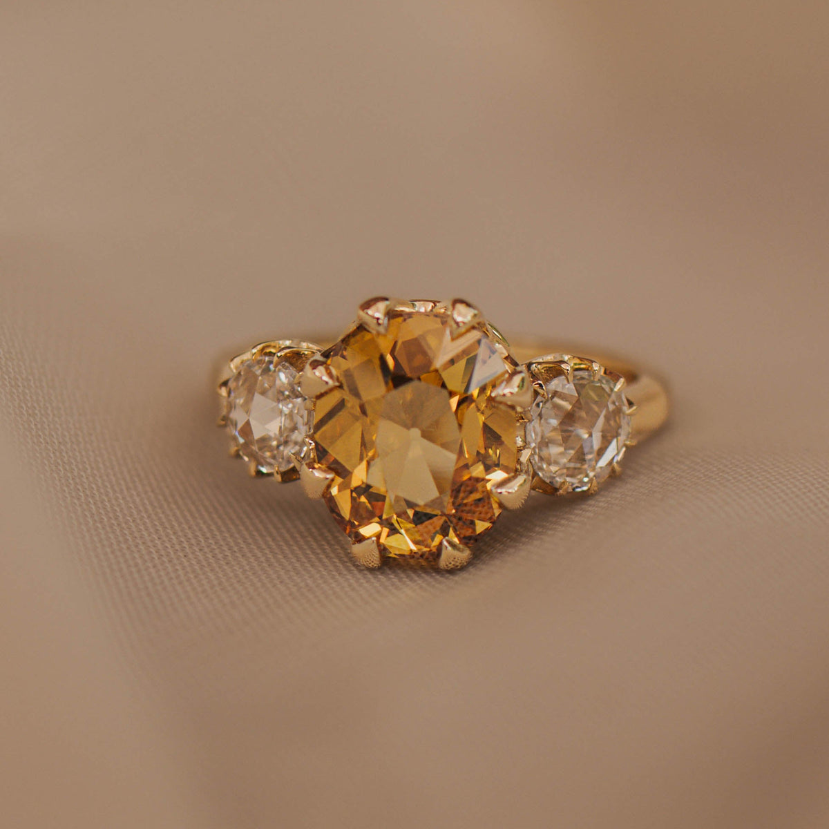A yellow gold ring featuring a large oval-cut yellow diamond in the center, flanked by two smaller round rose cut diamonds, resting on a soft beige fabric surface.