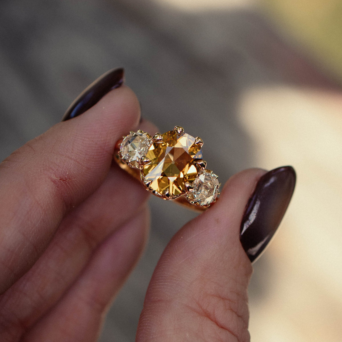 A gold ring with a large central yellow diamond and two smaller round rose cut diamonds, held between fingers against a blurred outdoor background.