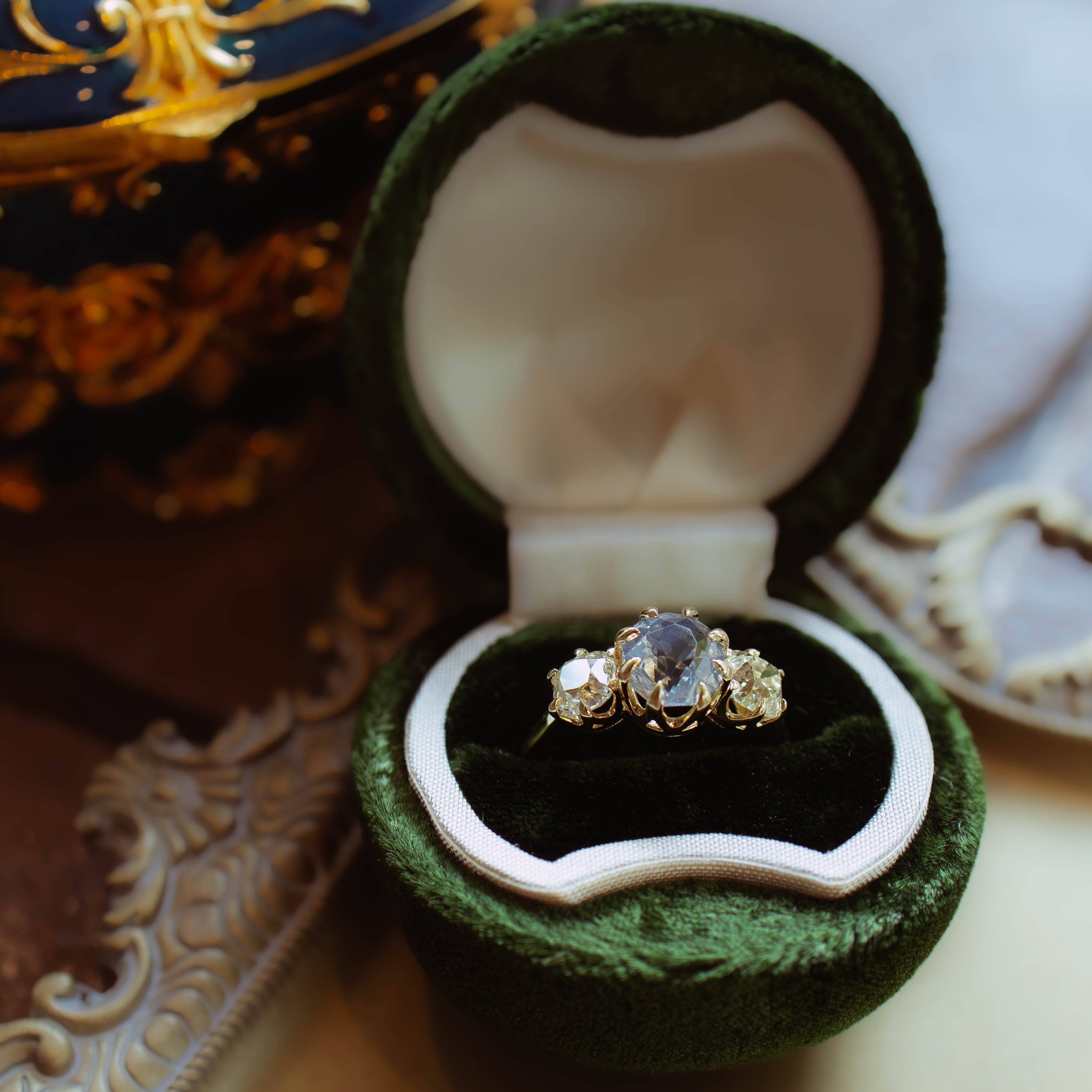 A gold ring with a central blue gemstone displayed in an open dark green velvet ring box, set against an elegant, softly blurred background with gold and blue decorative accents.