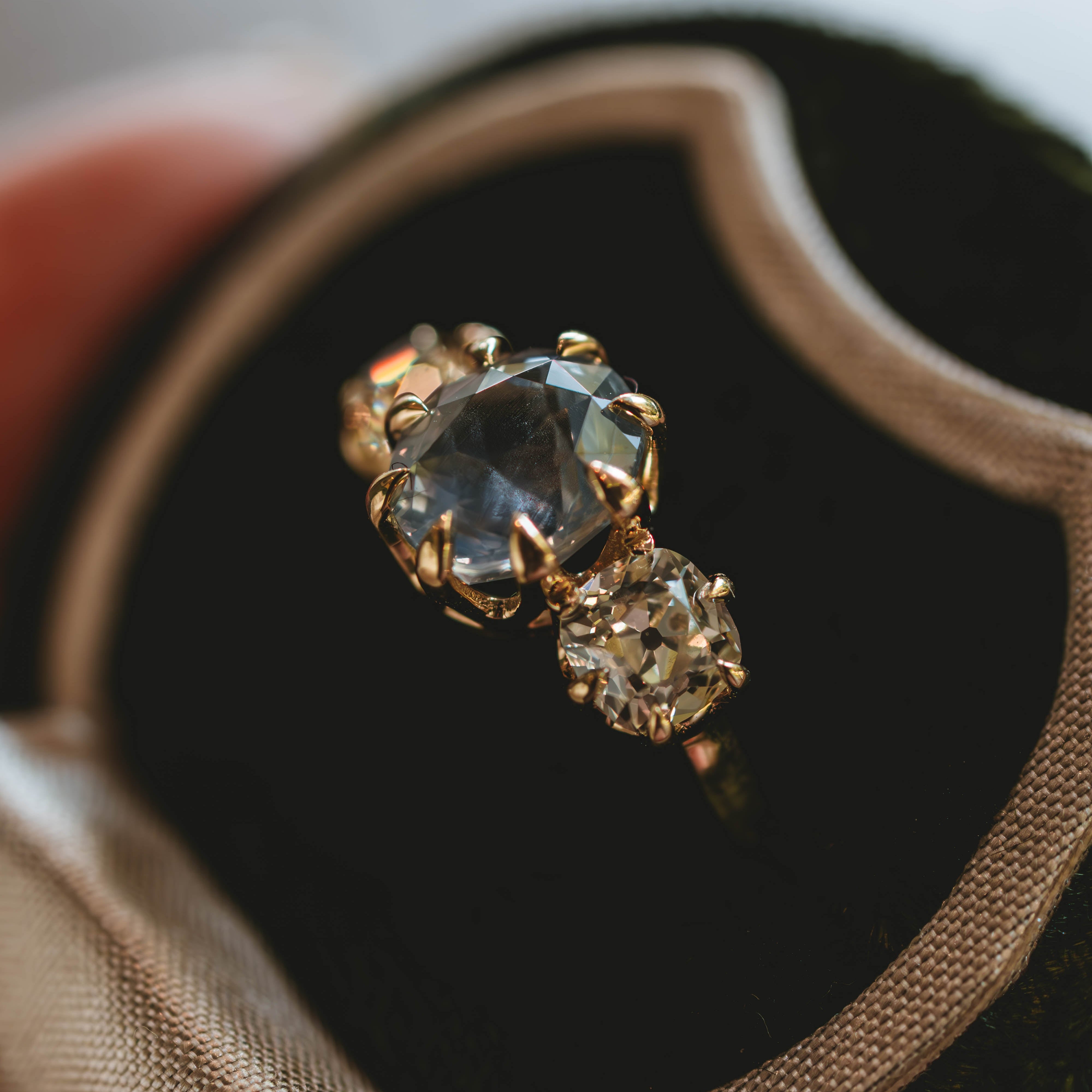 A close-up of a gold ring with a large oval sapphire emstone and a smaller cushion old mine diamonds, displayed inside a dark velvet ring box
