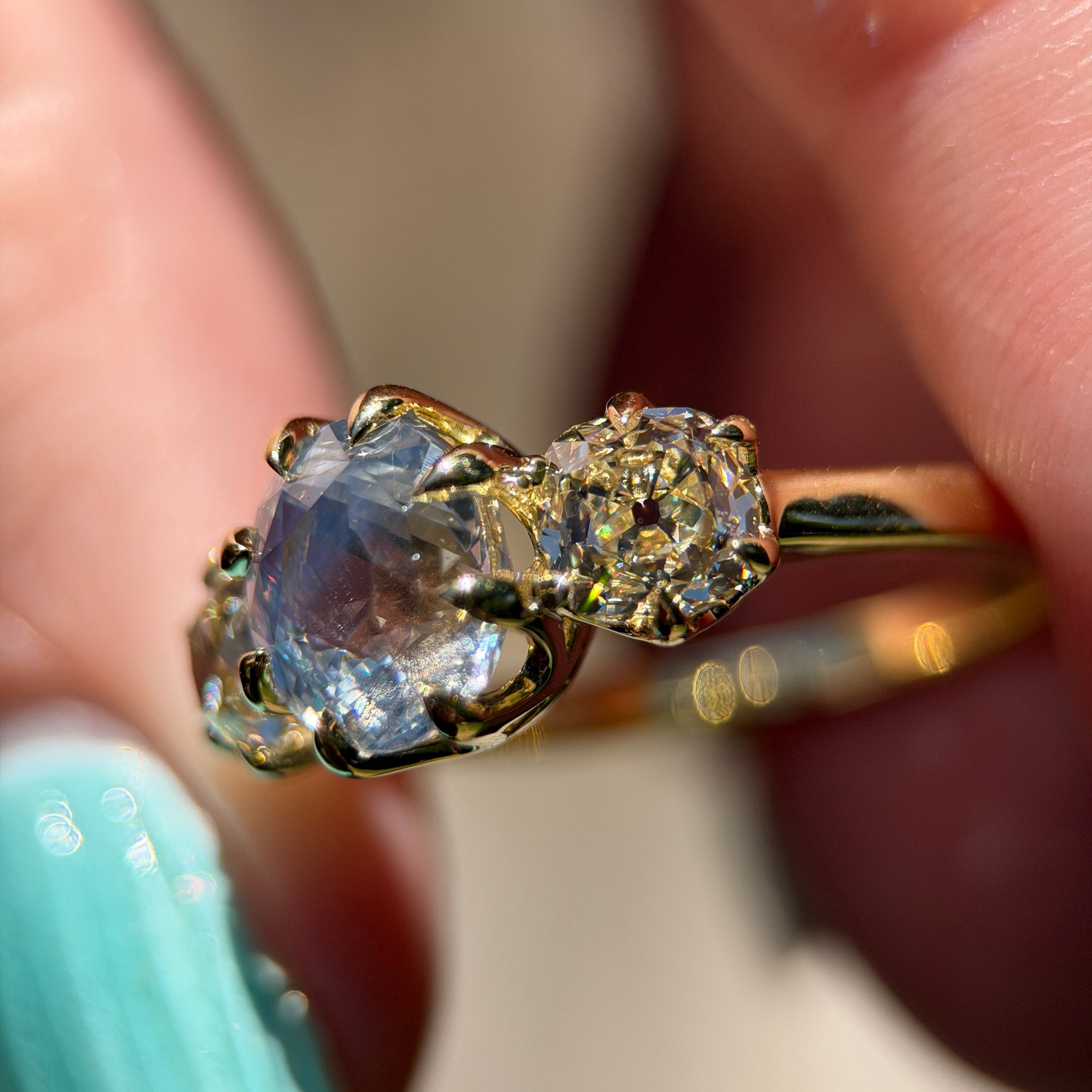A close-up of a gold ring with a large blue oval cut sapphire gemstone and smaller diamond accent stones, held between fingers against a soft blurred background.