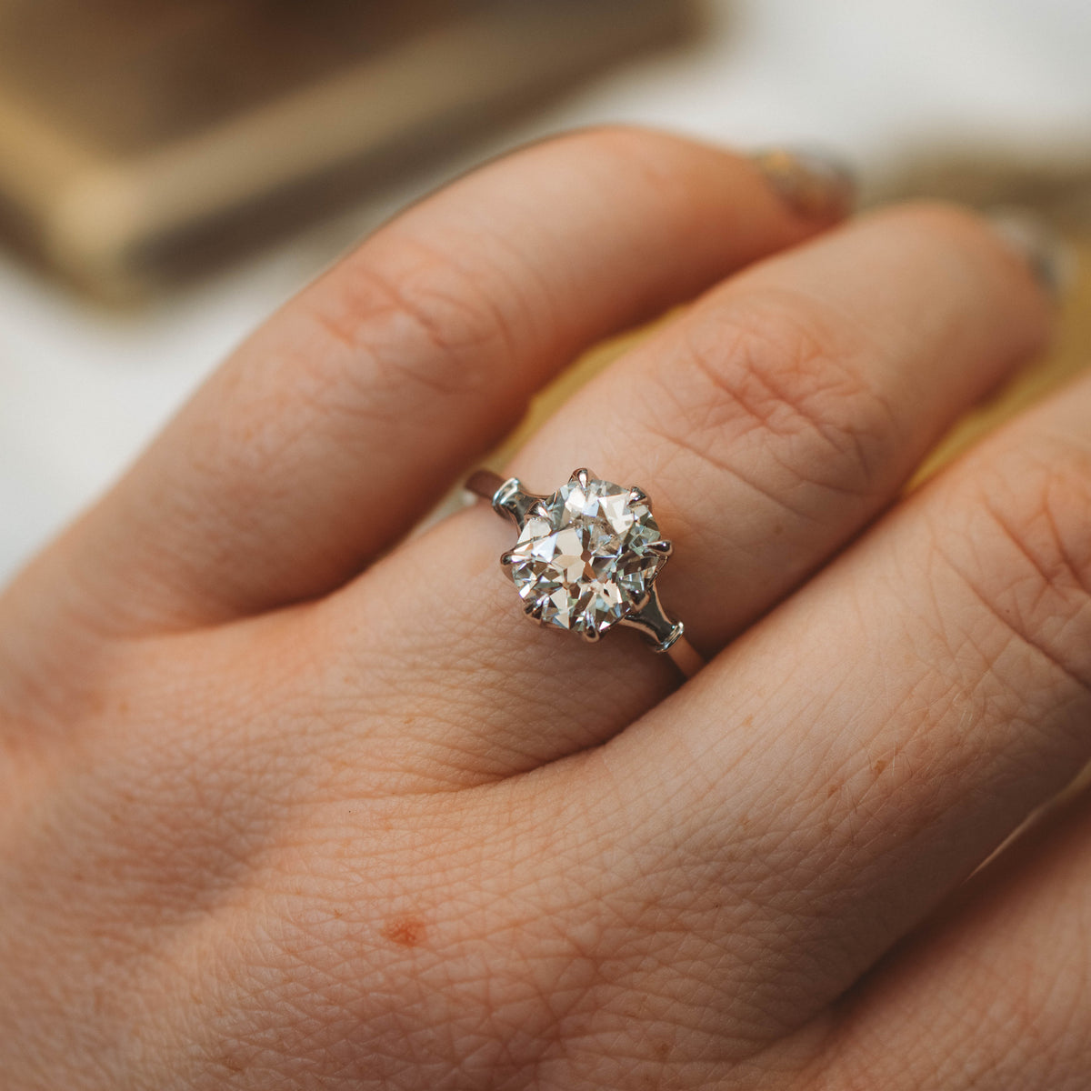 A close-up of a hand wearing a silver-toned ring with a cushion antique cut diamond. The ring is shown on the ring finger against a softly blurred background.