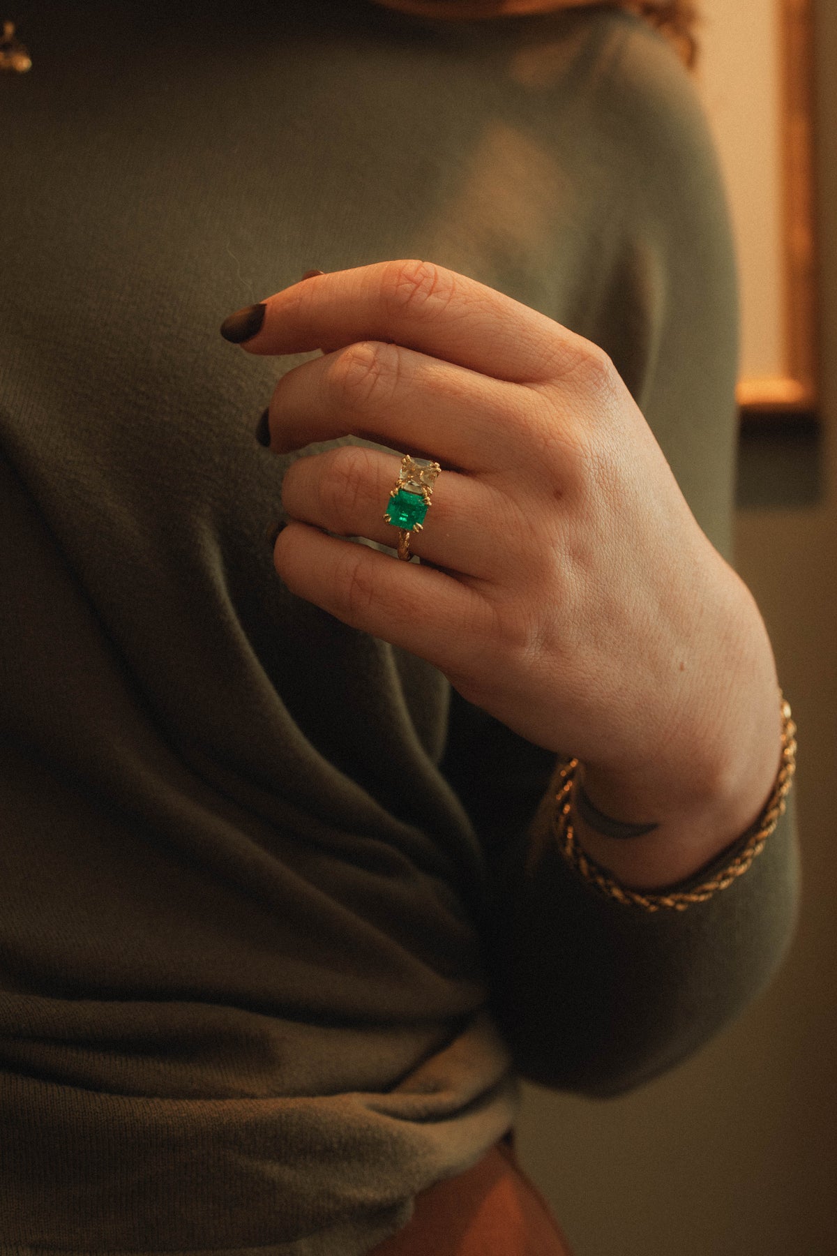A close-up of a person's hand wearing a gold ring with a square green emerald gemstone and cushion yellow diamond, paired with a gold chain bracelet, against a dark olive green top.